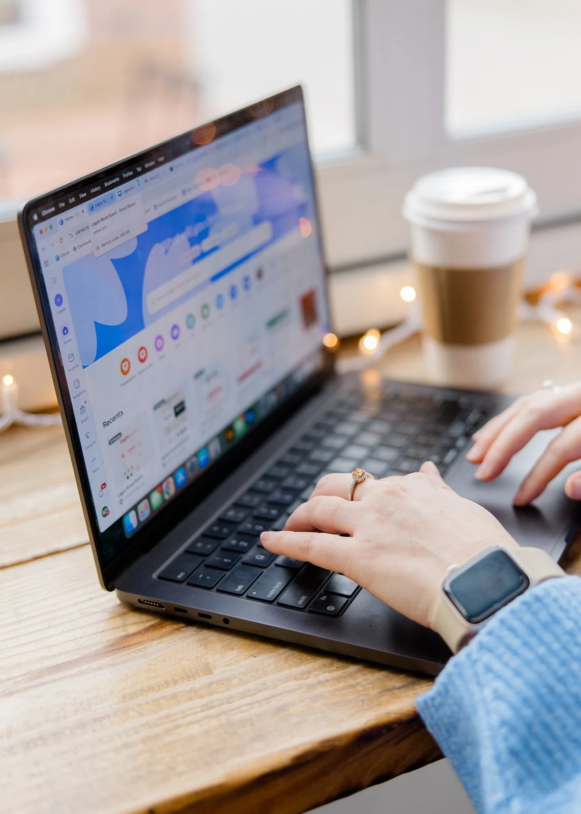 Person using a laptop with social media or website open, on a wooden table with a cup of coffee and a smartwatch visible.
