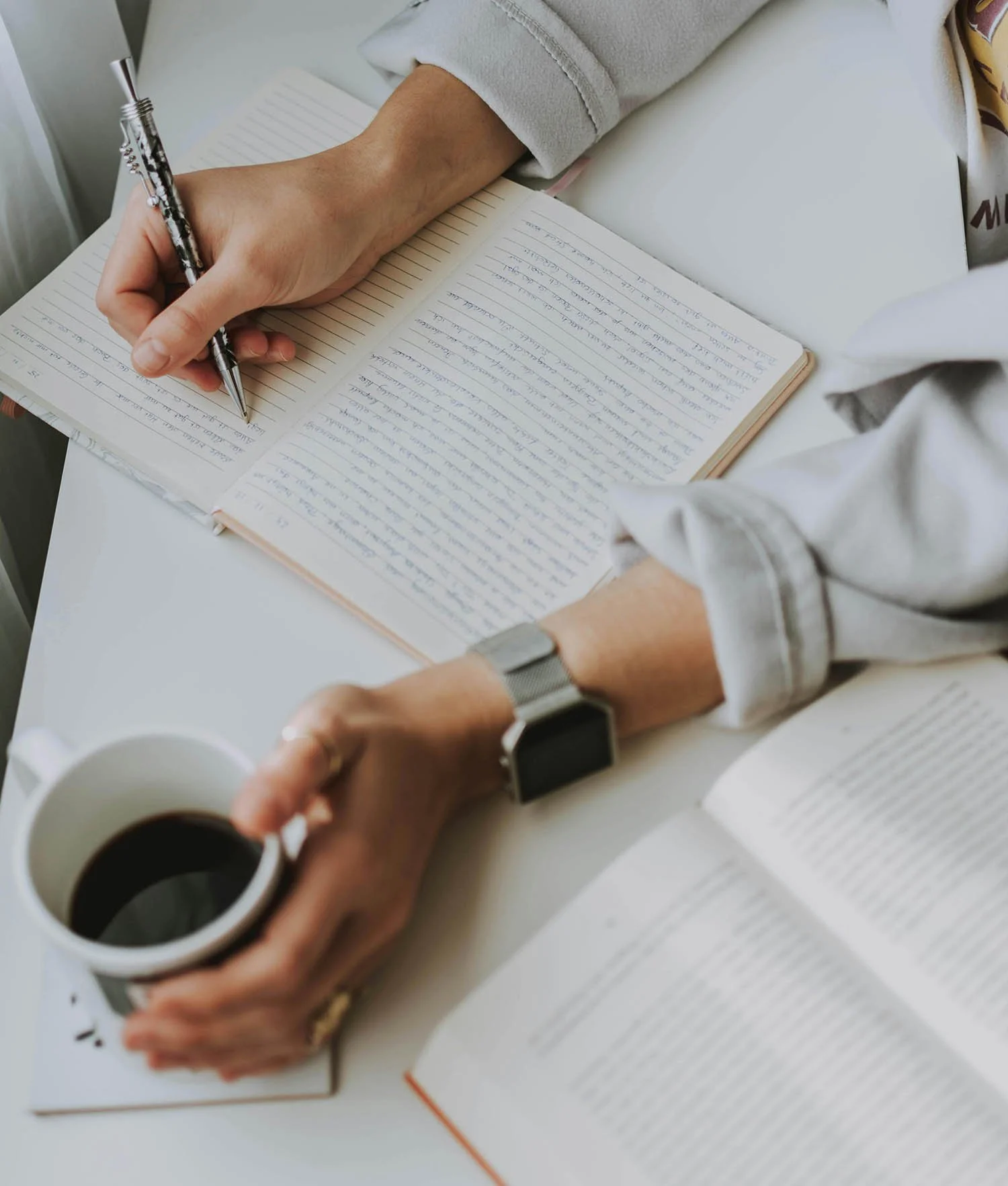 Person writing in a notebook with a pen, holding a cup of coffee, and surrounded by papers and books.