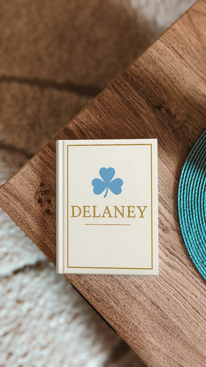 A beige book with gold lettering that reads 'DELANEY' and a blue four-leaf clover logo on a wooden table next to a teal woven placemat.