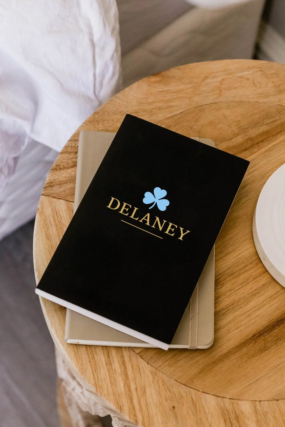 A black book with a blue three-leaf clover and the word 'DELANEY' in gold, resting on a beige book on a round wooden table.