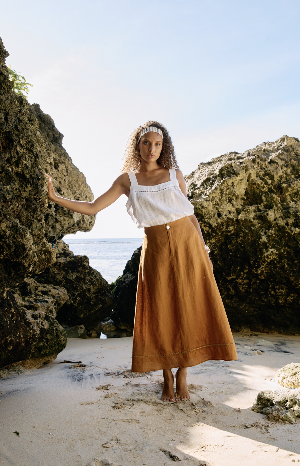 A woman standing on a sandy beach between large rocks, wearing a white sleeveless top, a long brown skirt, and a headband, with the ocean and sky in the background.