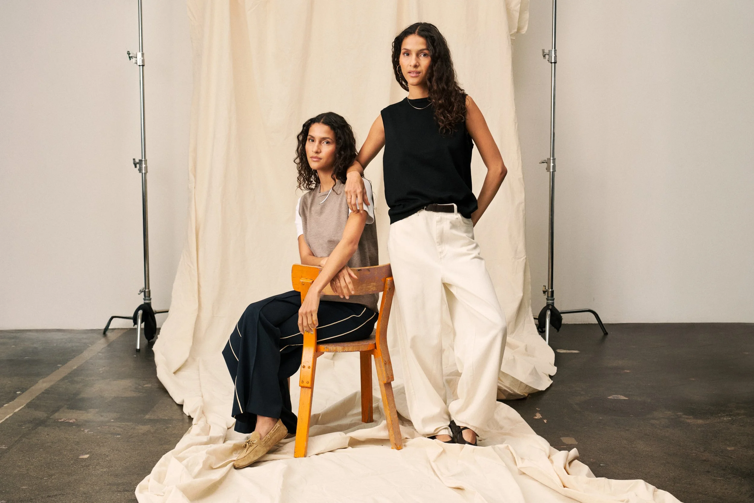 Two women with curly hair in a studio with a beige backdrop and lighting equipment. One woman is seated on a wooden chair, wearing a beige T-shirt and black pants with white stripes, and tan shoes. The other woman is standing, leaning on the seated woman with her arm on the back of the chair, wearing a black sleeveless top and white pants.