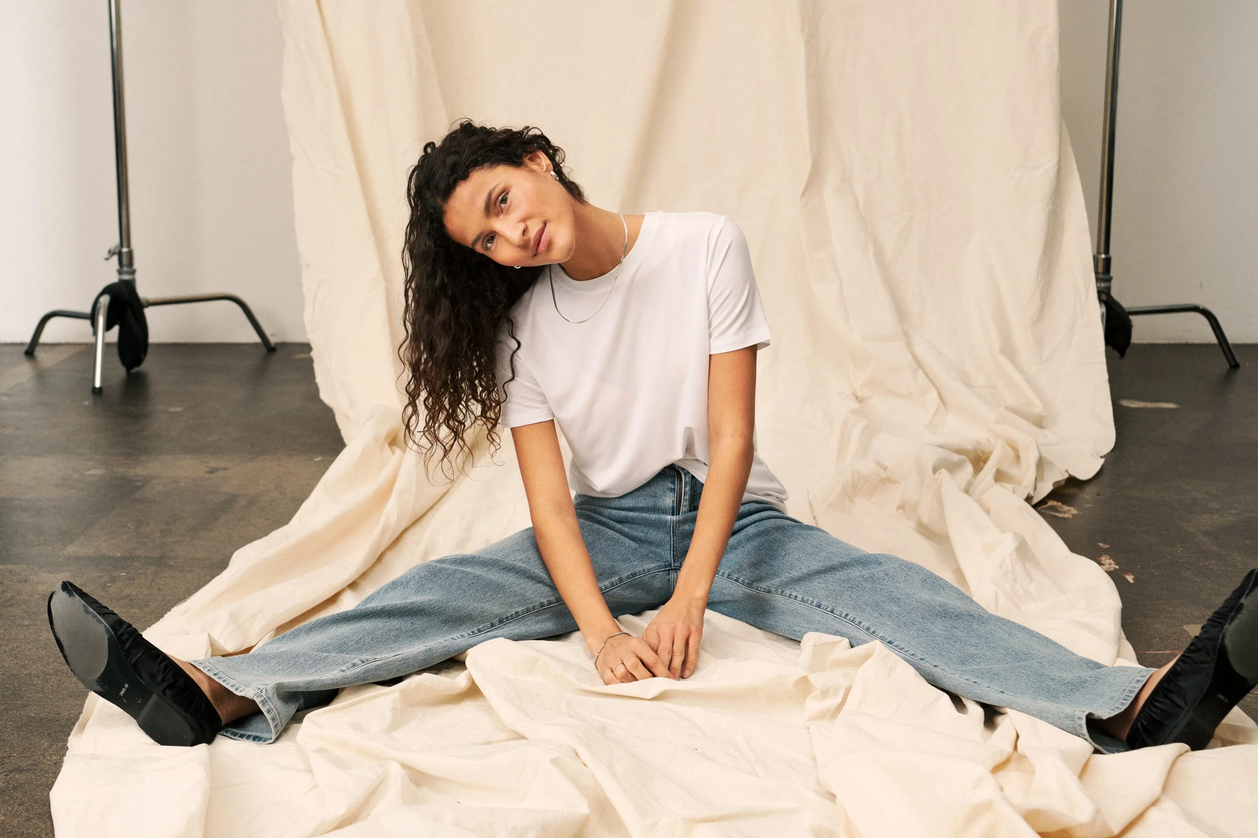 A young woman with curly dark hair, wearing a white t-shirt, blue jeans, and black slip-on shoes, sitting on a cream-colored fabric with legs spread apart, leaning her head to the side, in front of a cream-colored backdrop in a studio.