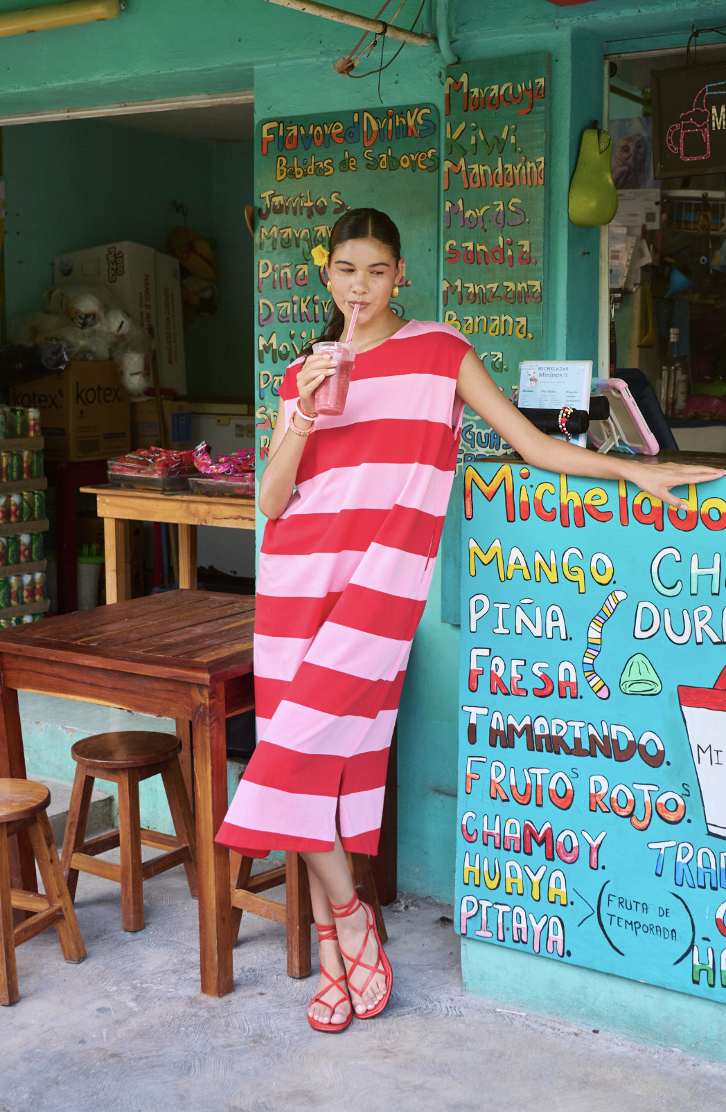Woman in a red and pink striped dress drinking a pink smoothie with a straw at a colorful fruit stand.