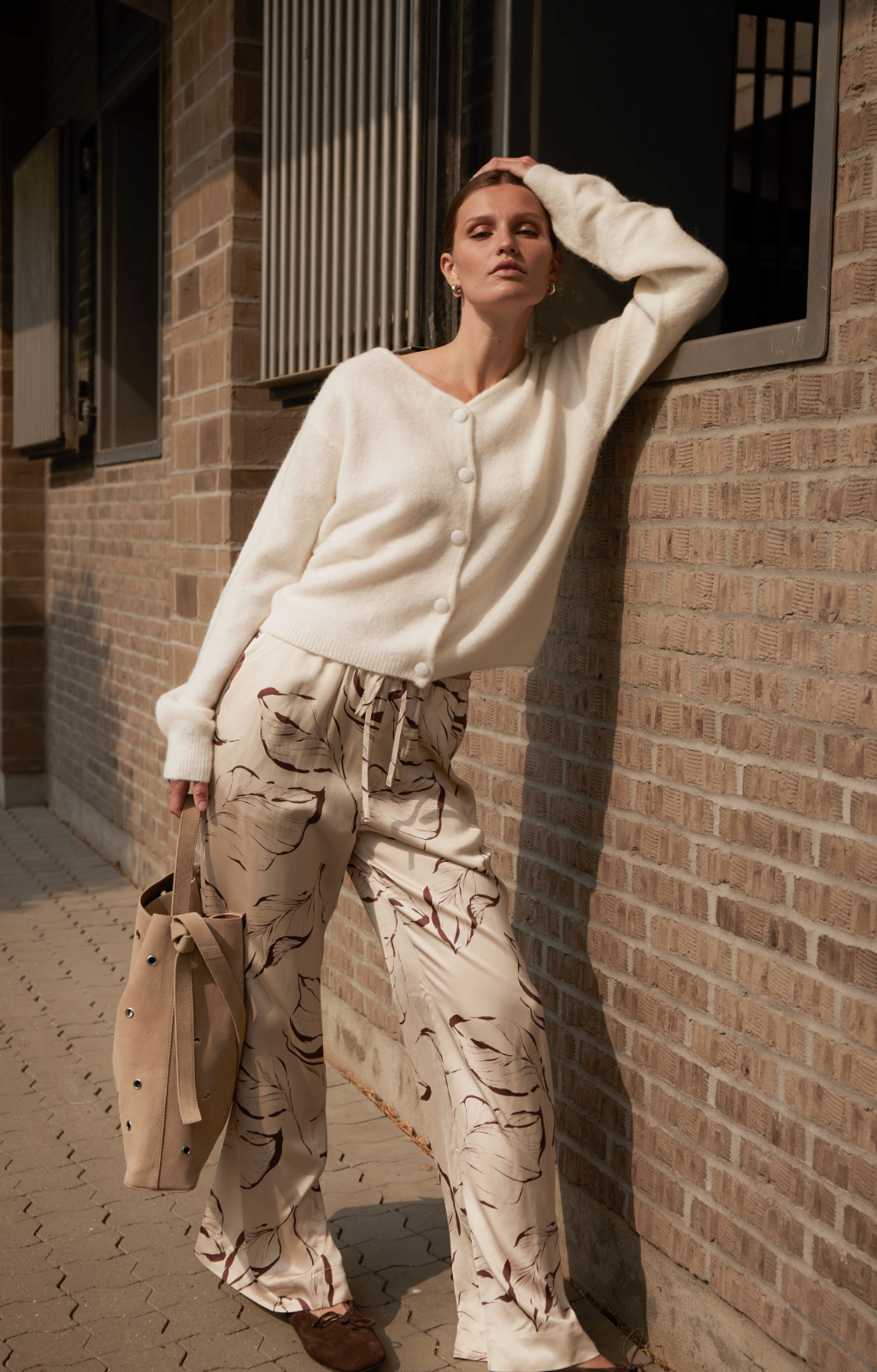 A woman leaning against a brick wall, wearing a cream-colored cardigan and wide-leg pants with a leaf pattern, holding a beige tote bag.