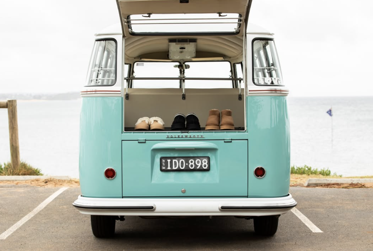 Rear view of a vintage turquoise and white Volkswagen van with an open back showing three pairs of shoes on the inside ledge, parked in a coastal area near water with a flag on a pole.