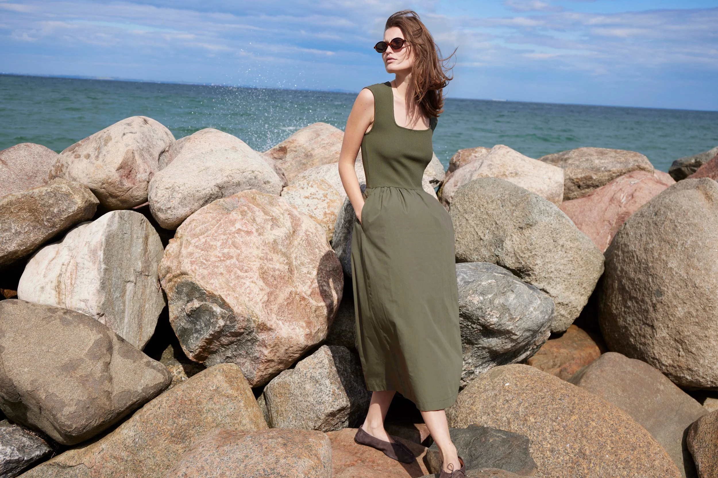 A woman in an olive green dress and sunglasses standing on rocks by the ocean on a partly cloudy day.