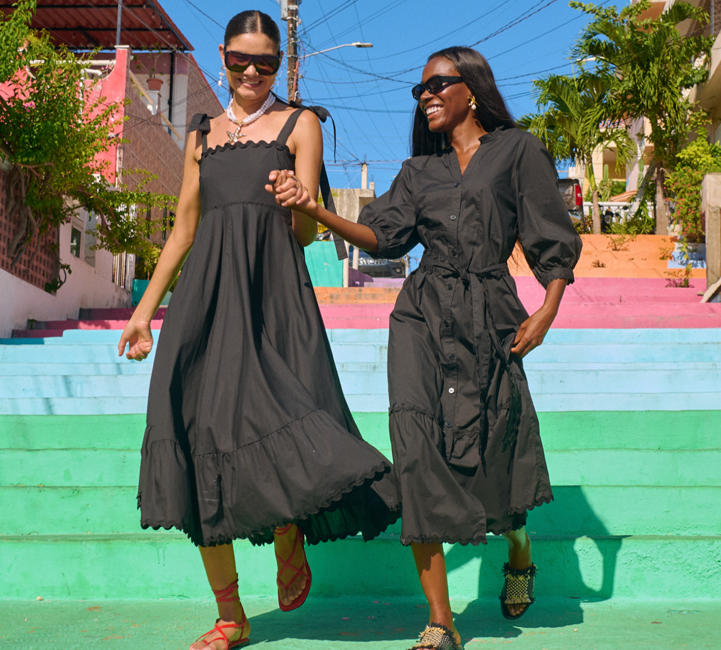 Two women with dark hair and sunglasses, wearing black dresses, dancing on colorful stairs outdoors under clear blue skies, with trees and buildings in the background.