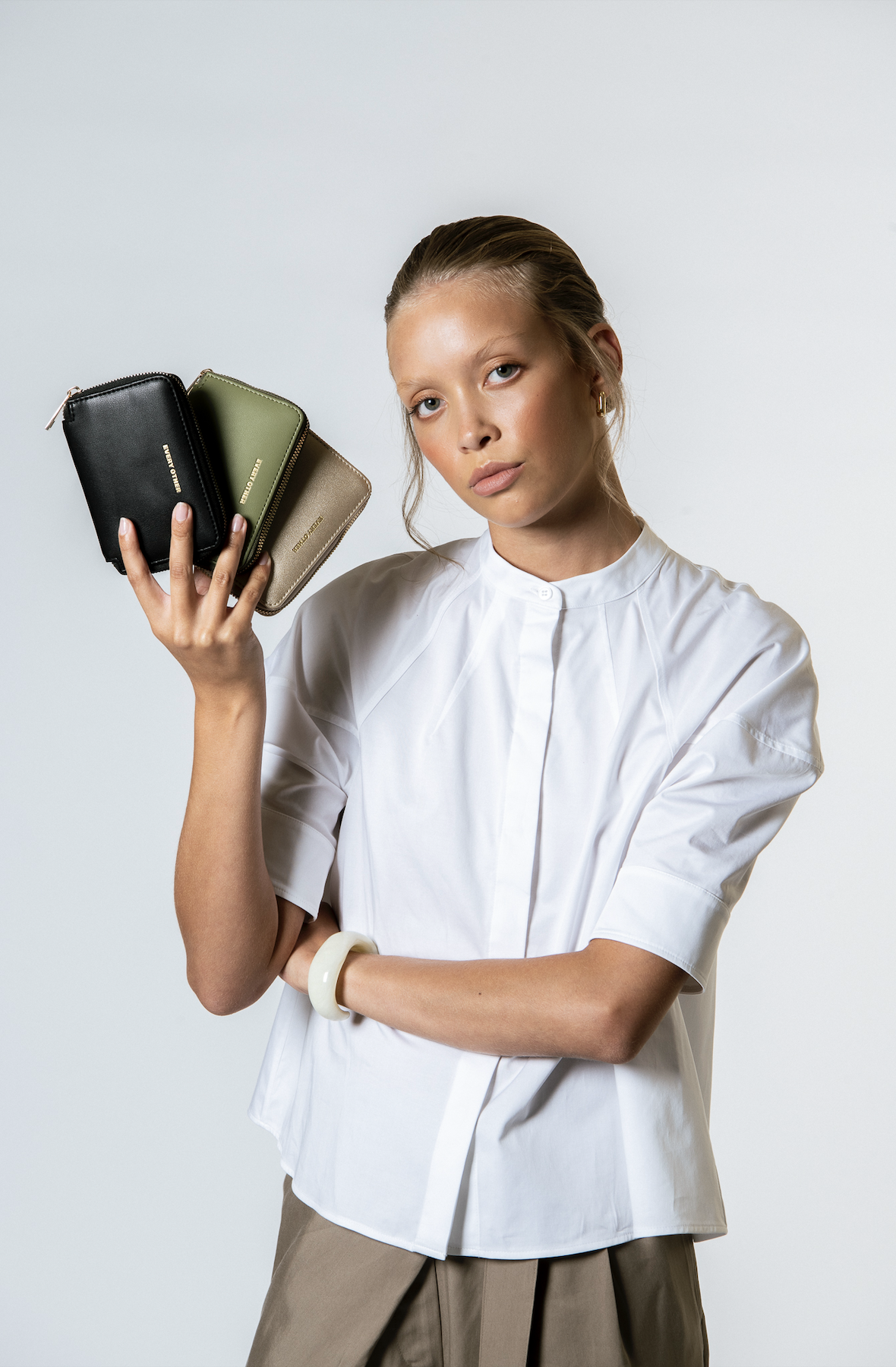 A woman with blonde hair tied back, wearing a white blouse with a high collar and beige pants, holds three small handbags in black, green, and beige in her right hand, against a plain light background.