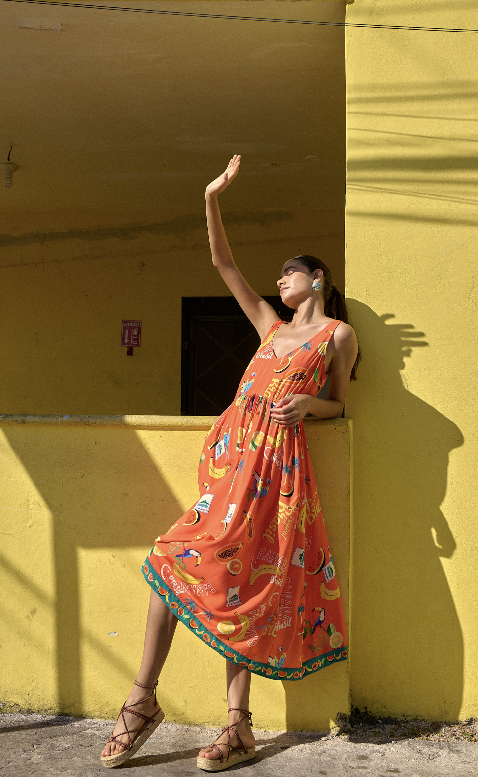 A woman in a colorful sundress stands against a yellow wall, raising her right arm while tilting her head back softly, in bright sunlight.