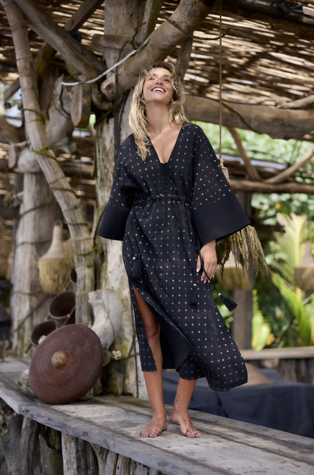 A woman wearing a black dress with white pattern, standing on a wooden platform outdoors, smiling and looking up, surrounded by a rustic tree and wooden structure.