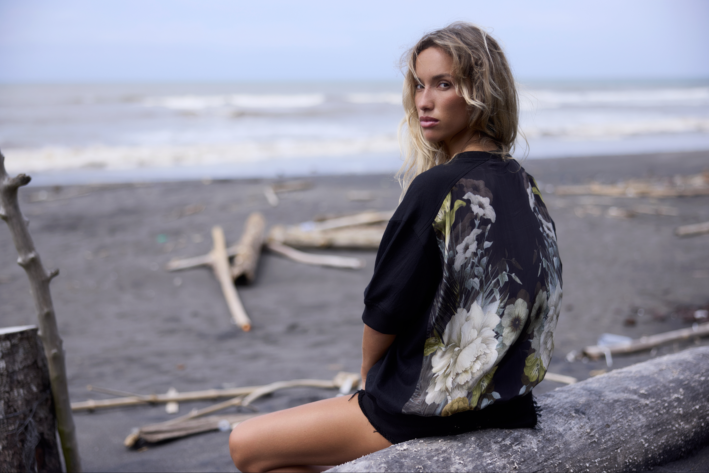 A young woman with wavy blonde hair sitting on a fallen log on a beach, facing sideways but looking at the camera, with the ocean waves in the background.