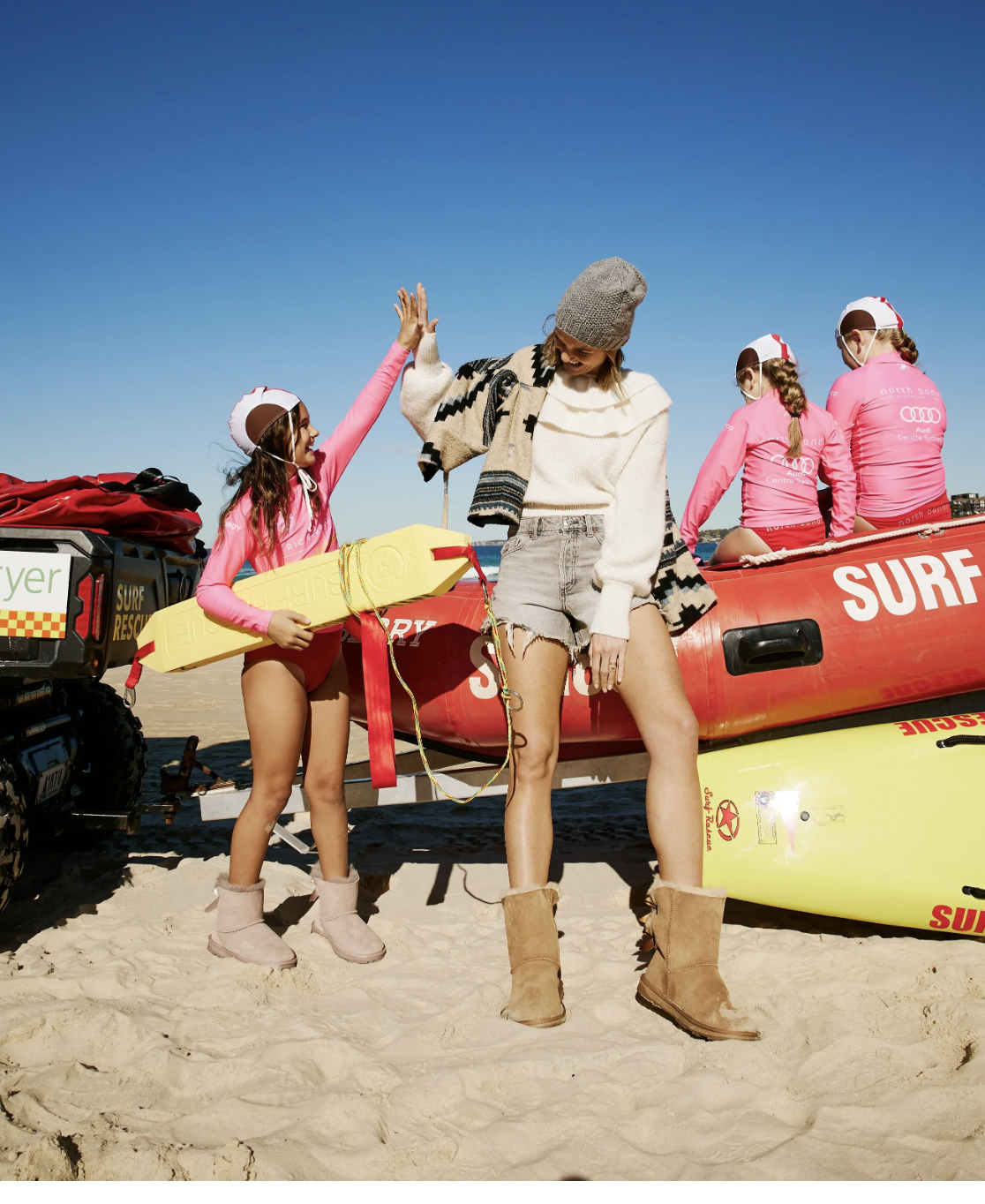 A woman and girl high-five on a sandy beach with a rescue boat and kids in pink surf gear in the background.