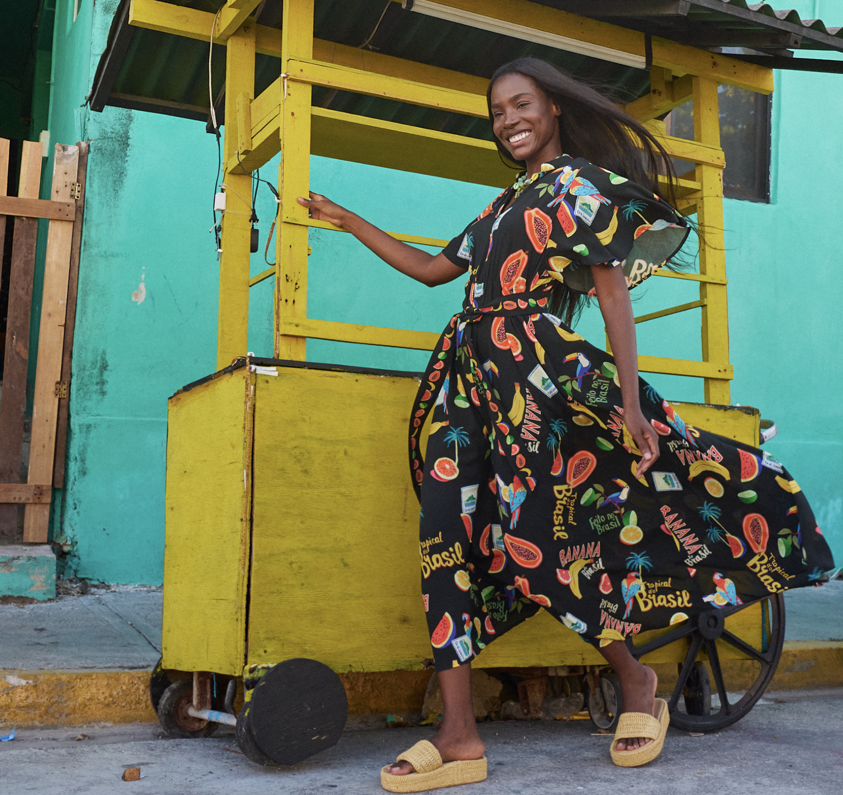 A woman with long hair, wearing a colorful dress with tropical fruit patterns, smiling while standing next to a yellow fruit stand with wheels outside a turquoise-colored building.
