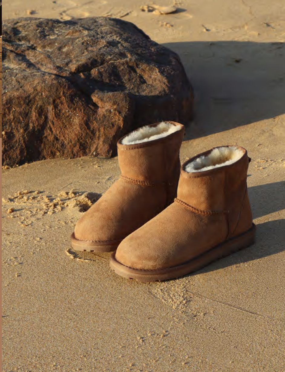 Pair of tan, sheepskin-lined winter boots on sandy beach with large rock in background.