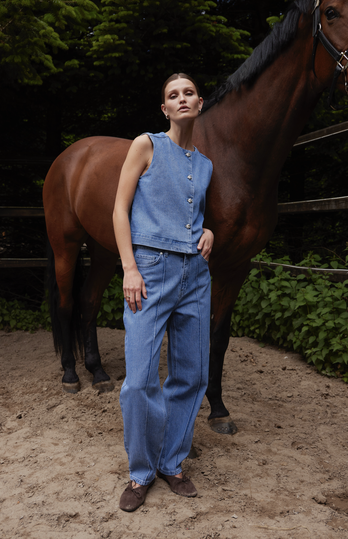 A woman in a sleeveless denim vest and high-waisted jeans stands in front of a brown horse, outdoors on a dirt surface with green foliage and a wooden fence in the background.