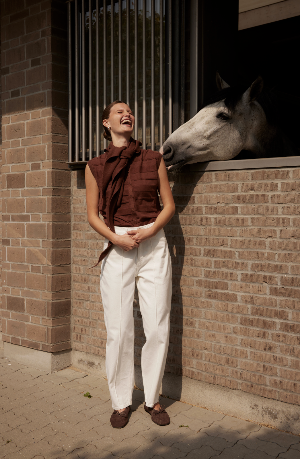 A woman laughing with her hands clasped in front of her, standing next to a gray horse that is leaning its head over a brick stable wall.