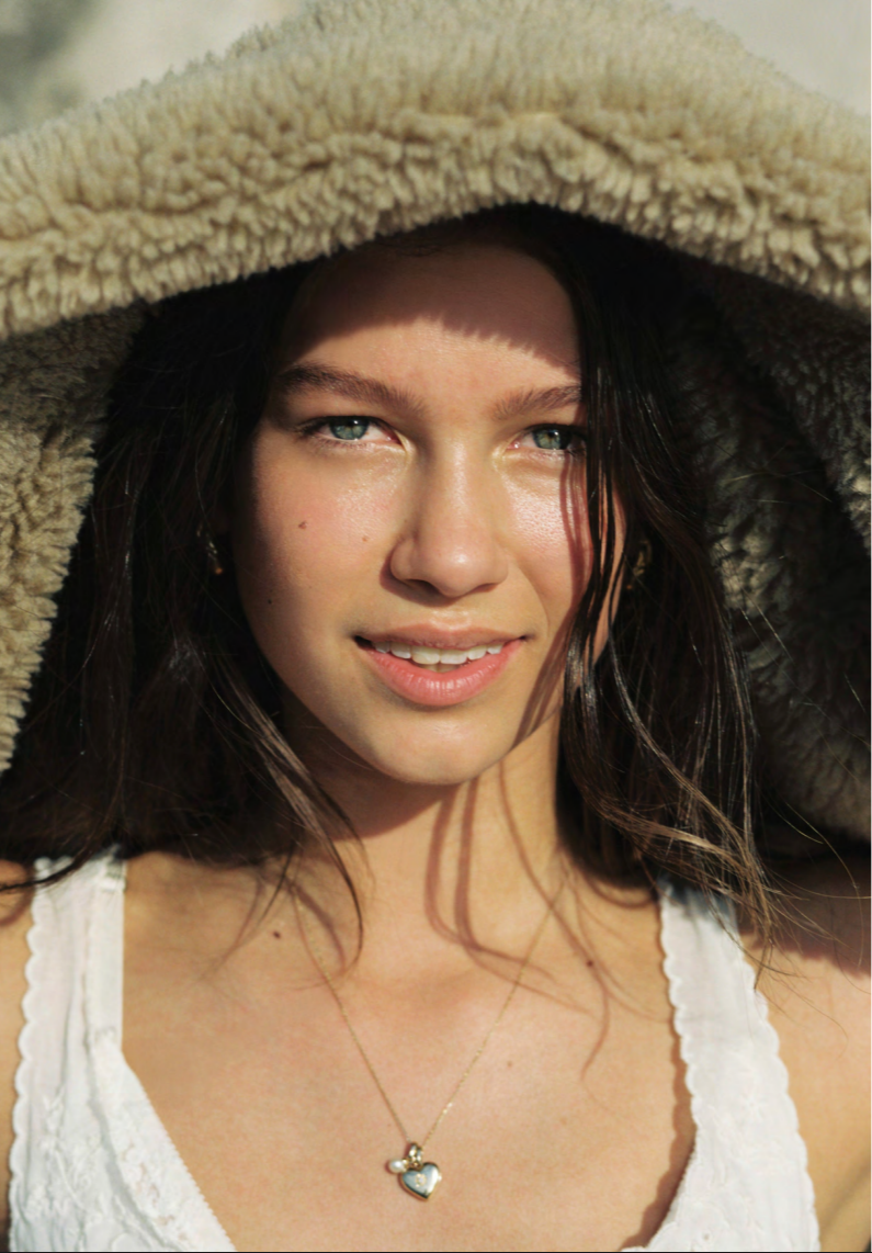 Close-up of a woman with blue eyes and dark hair, smiling under a large, furry beige hood, wearing a white top and a gold necklace with a heart-shaped pendant.