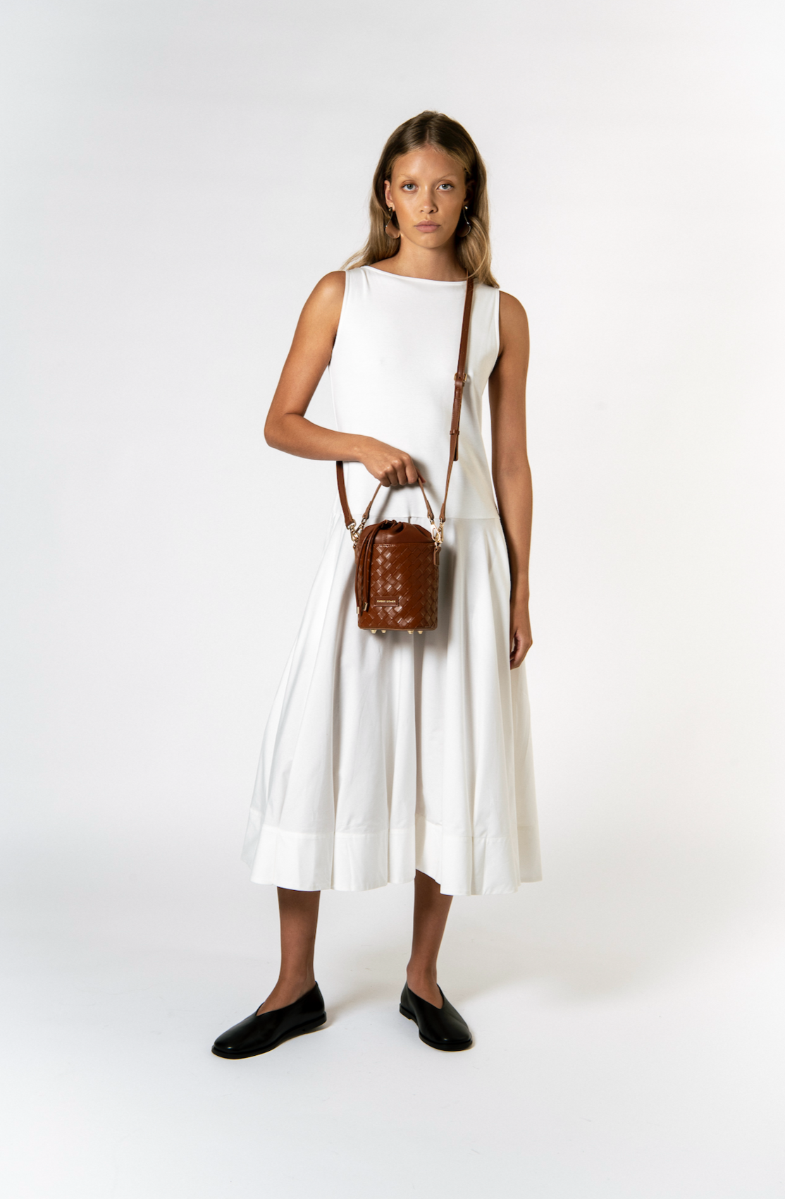 A young woman wearing a white sleeveless dress, black shoes, and carrying a small brown woven handbag.