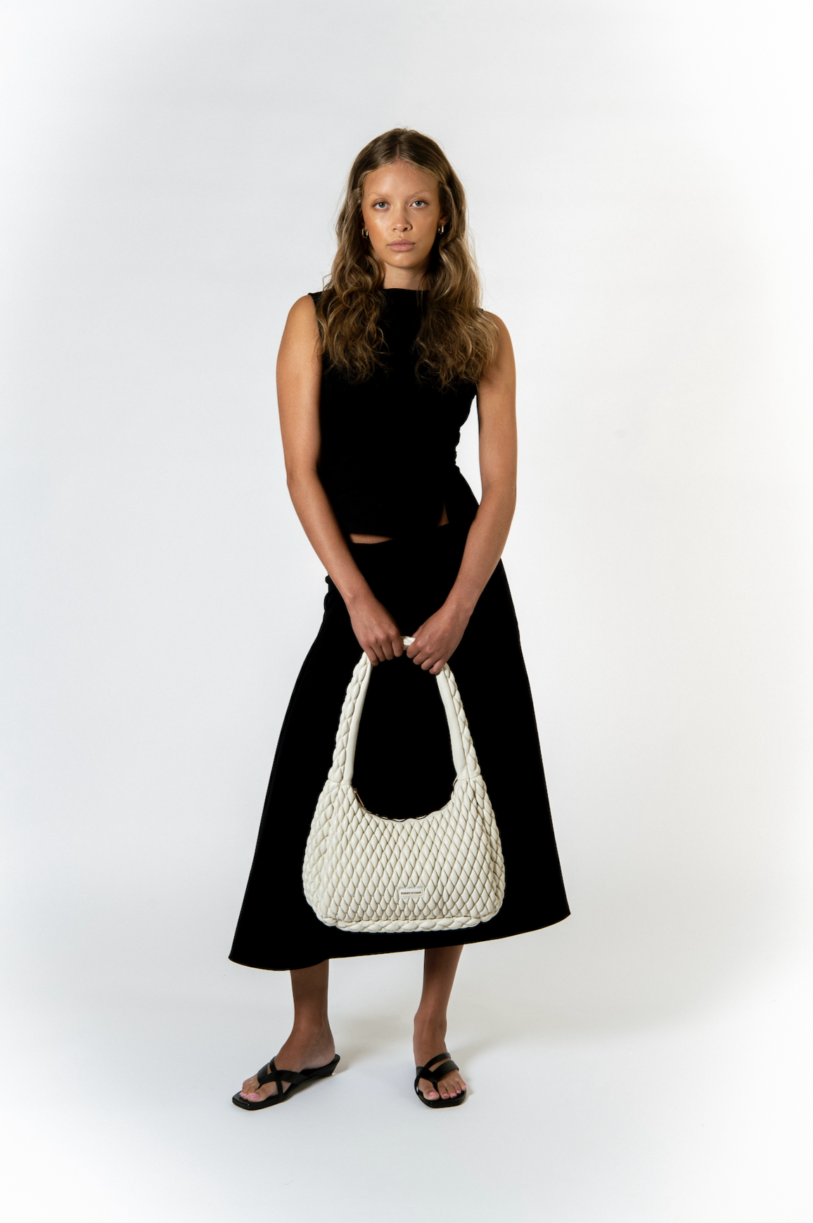 A young woman with wavy brown hair wearing a sleeveless black top, black skirt, black sandals, and holding a large cream-colored, woven handbag against a plain white background.