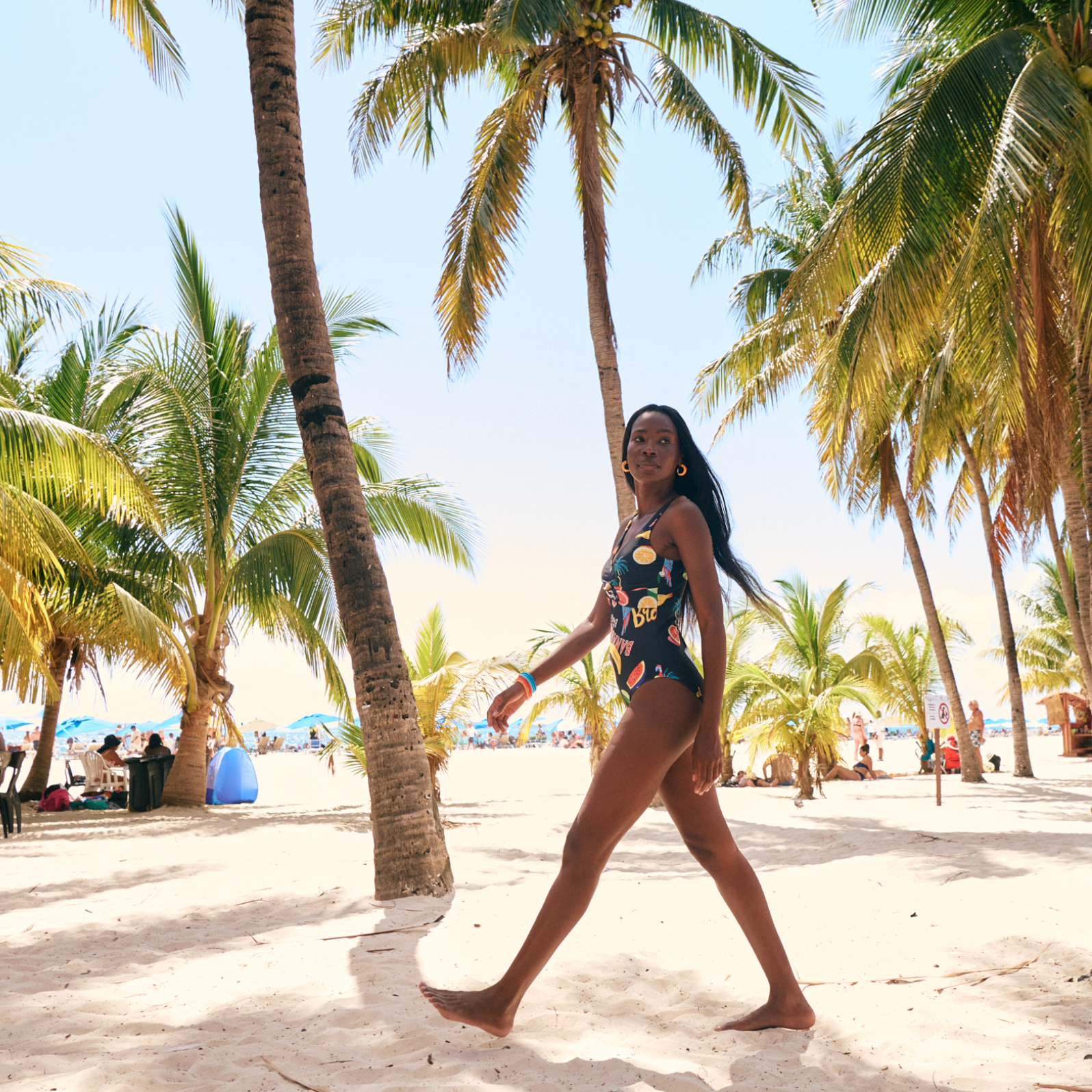 A woman in a colorful swimsuit walking barefoot on a sandy beach with palm trees and umbrellas in the background.