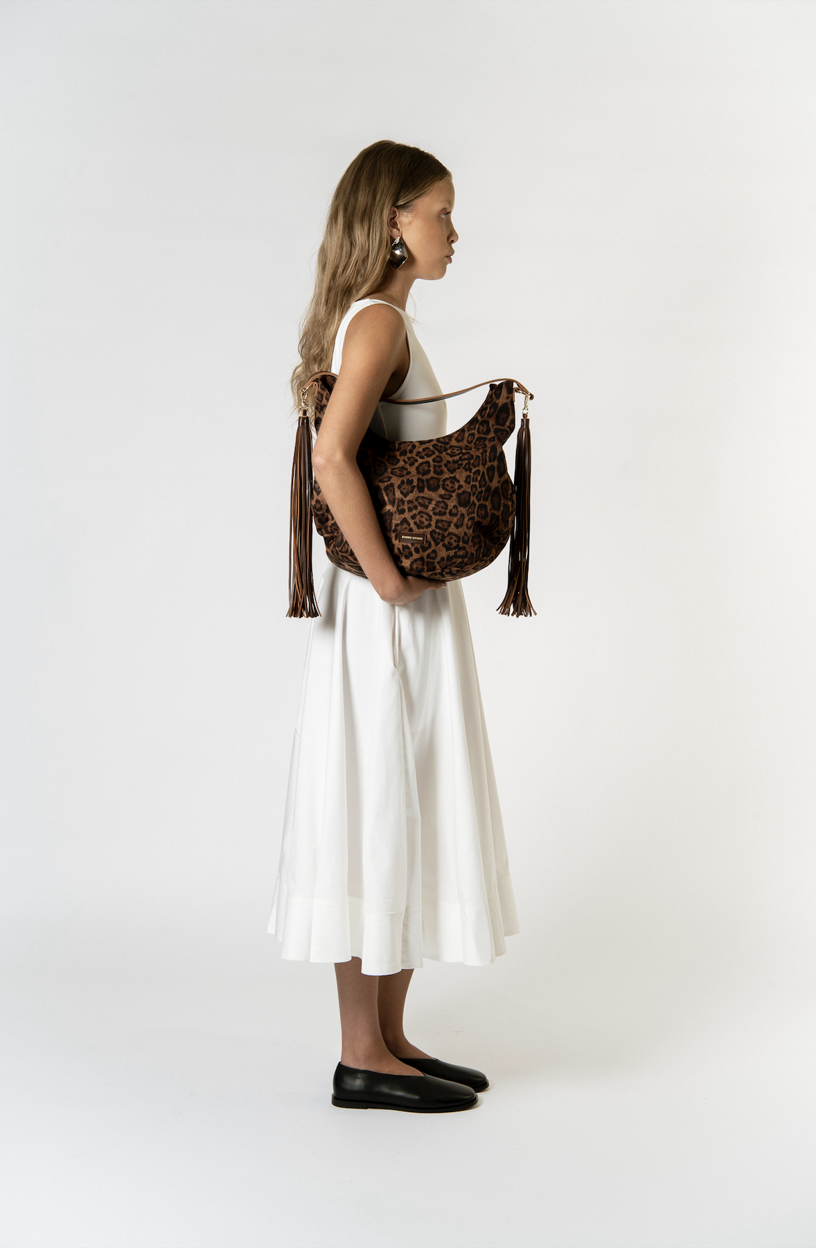 A woman dressed in a white sleeveless dress and black shoes, holding a leopard print bag with tassels.