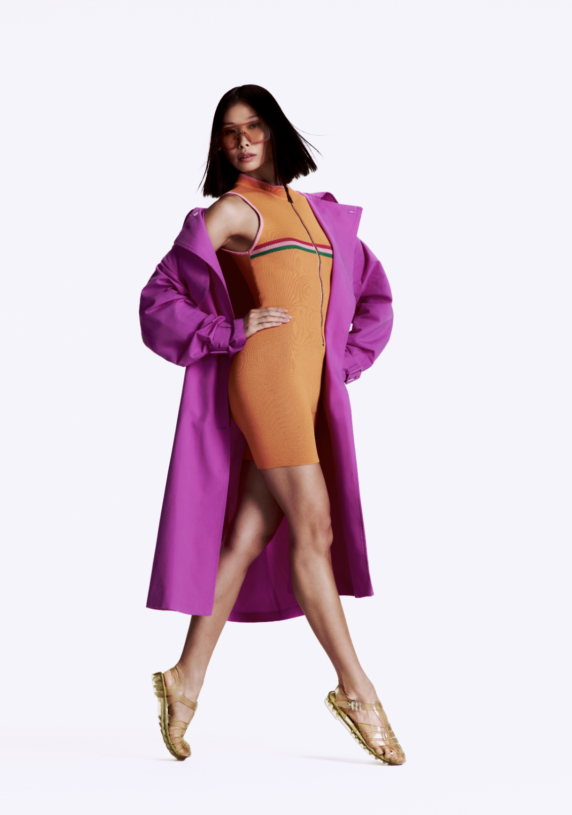 A woman wearing sunglasses, a sleeveless orange dress with green and pink stripes, a purple trench coat, and gold shoes, posing confidently against a plain white background.