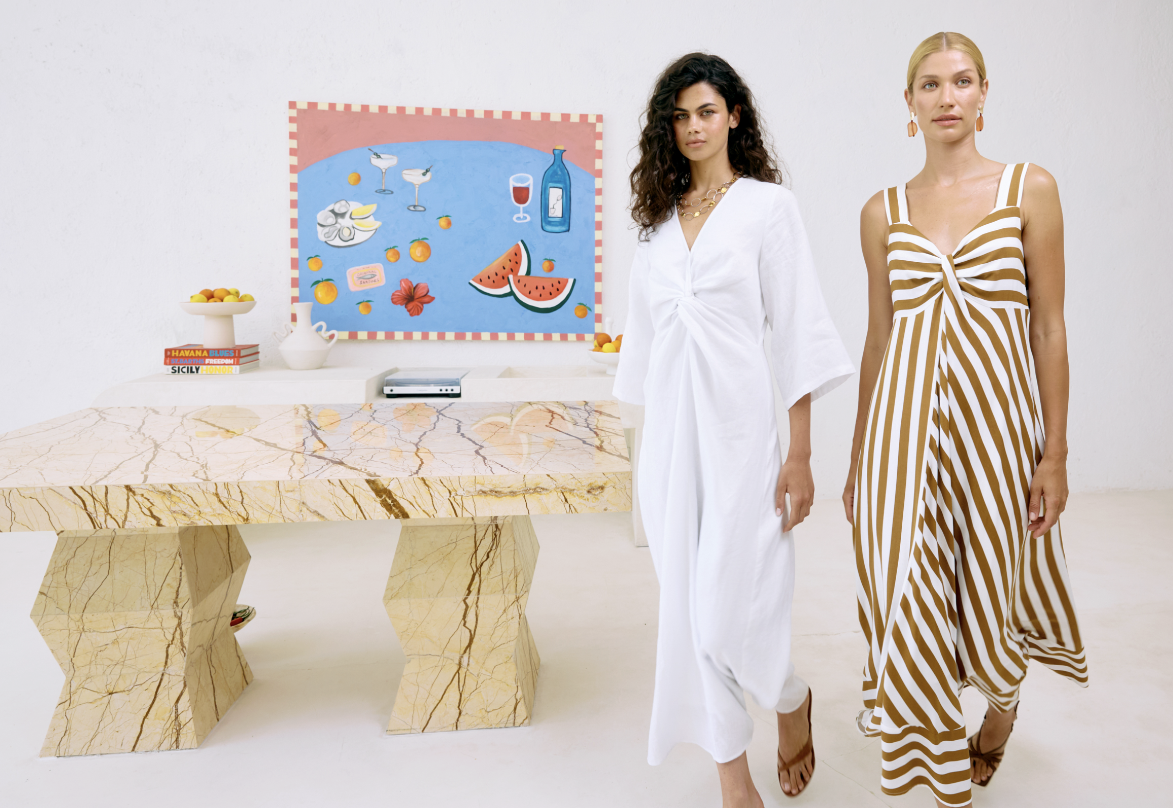 Two women walking past a marble table in an art gallery or studio. One woman is wearing a white dress, the other is wearing a brown and white striped dress. There is a colorful painting of fruit and drinks on the wall behind them, along with a bowl of fruit and books on the table.