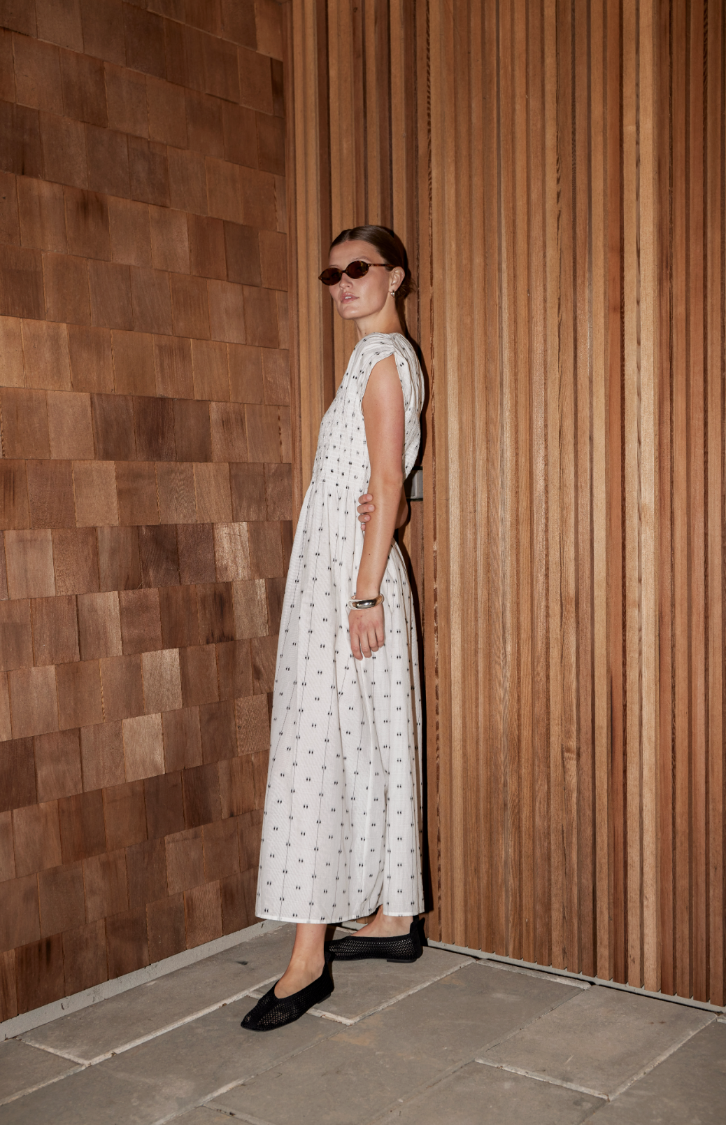 A woman wearing a white dotted maxi dress, black shoes, and sunglasses standing against a two-tone wooden wall.