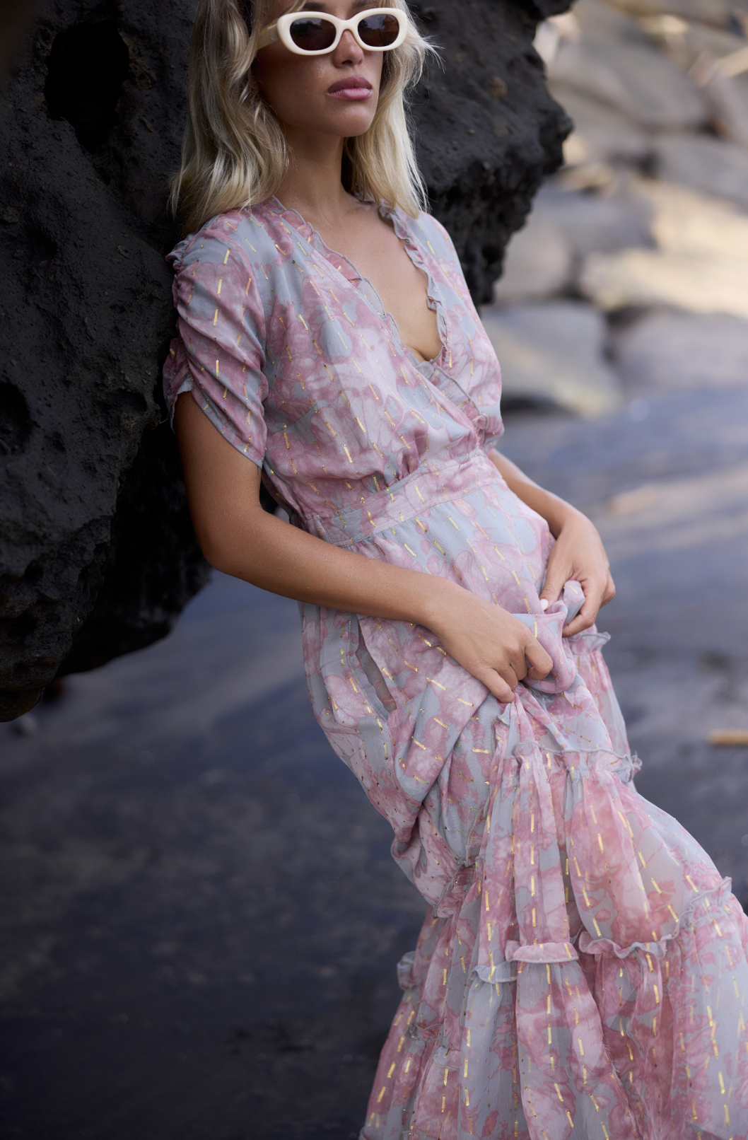 A woman with long blonde hair wearing white sunglasses and a pink ruffled dress with gold accents, standing outdoors near rocks and water.