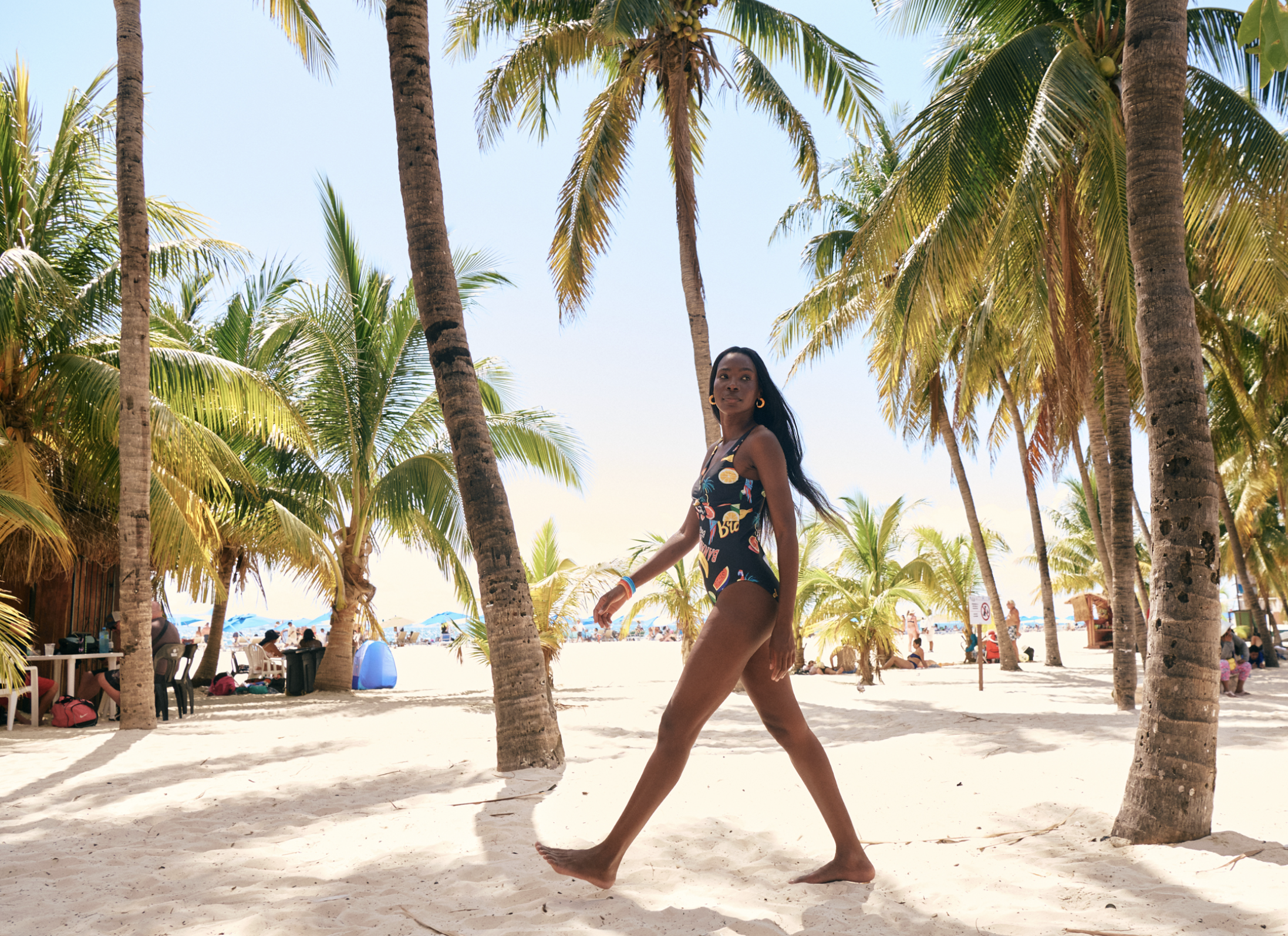 A woman in a black swimsuit with colorful patterns walking barefoot on a sandy beach surrounded by tall palm trees, with people and beach umbrellas in the background on a sunny day.
