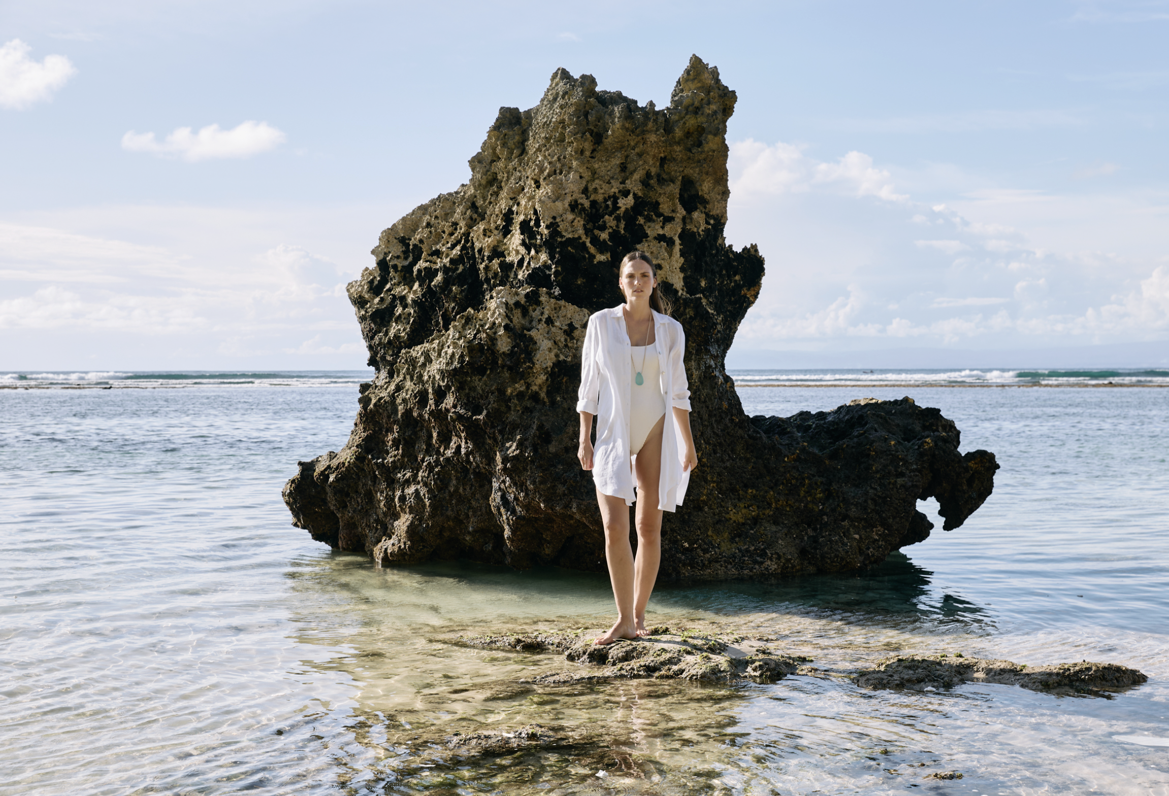 Woman in white dress and open white shirt standing on rocks in shallow sea with large rock formation behind her, under a partly cloudy sky.