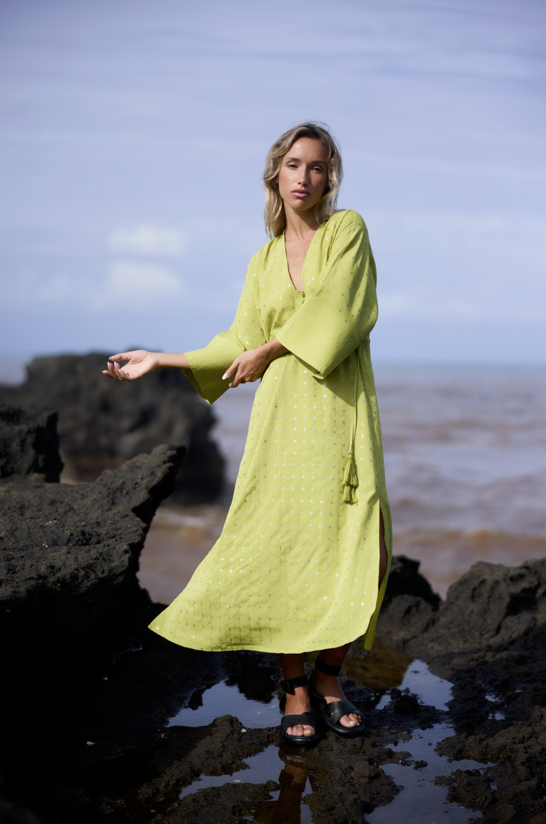 A woman stands on black rocks by the ocean wearing a yellow-green dress and black sandals, with a cloudy sky in the background.