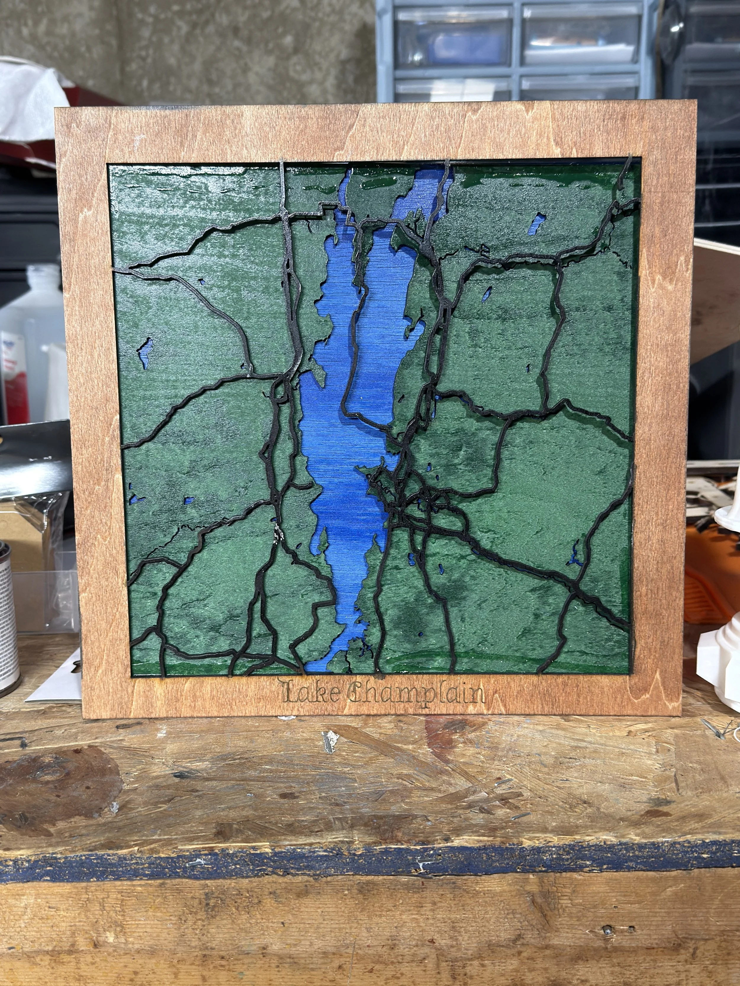 A stained glass artwork of Lake Champlain with green landscape and blue water, framed with wood, placed on a wooden table.