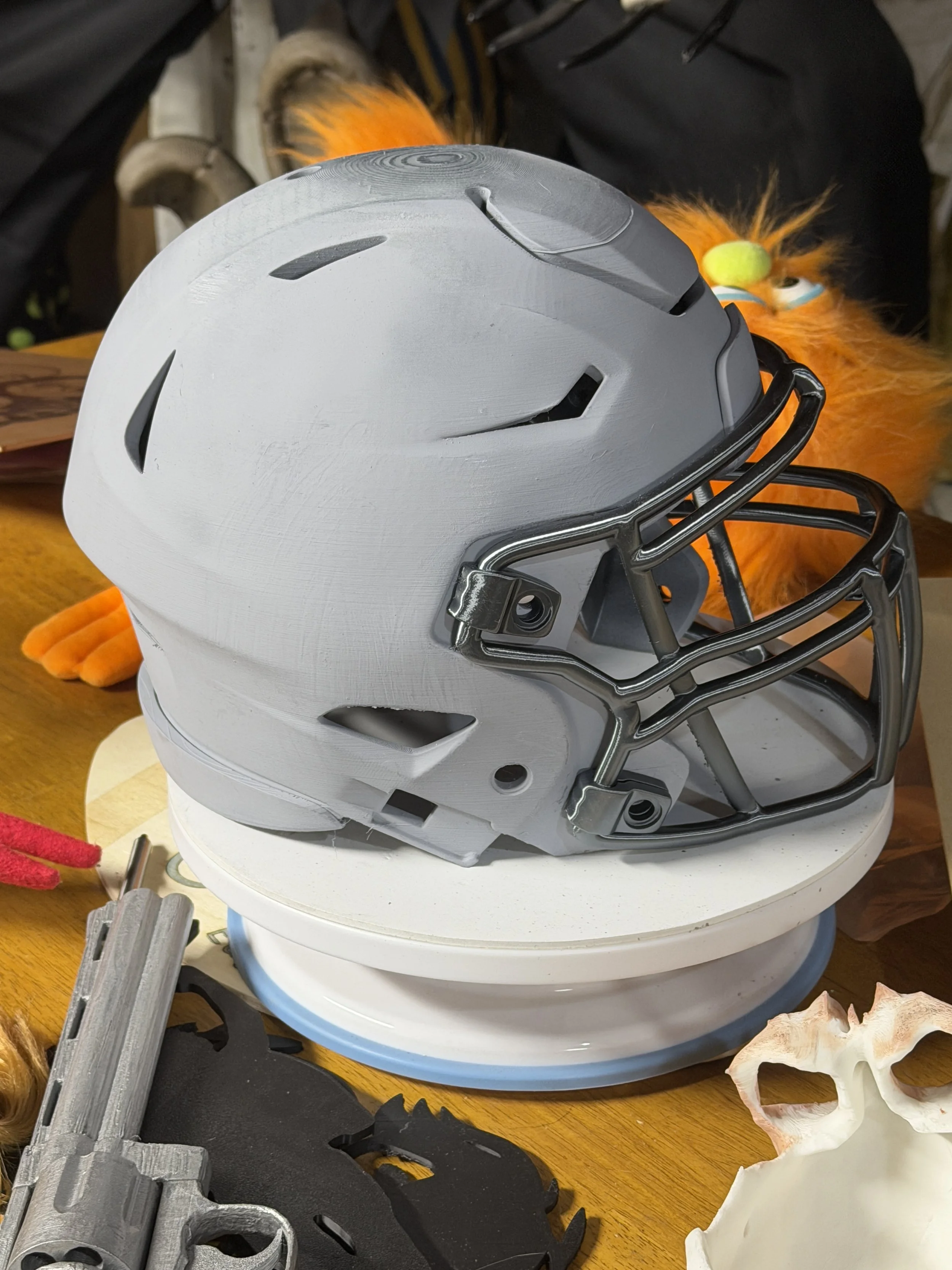 Gray football helmet resting on a white platform on a wooden table, with Halloween decorations and a toy on the table.