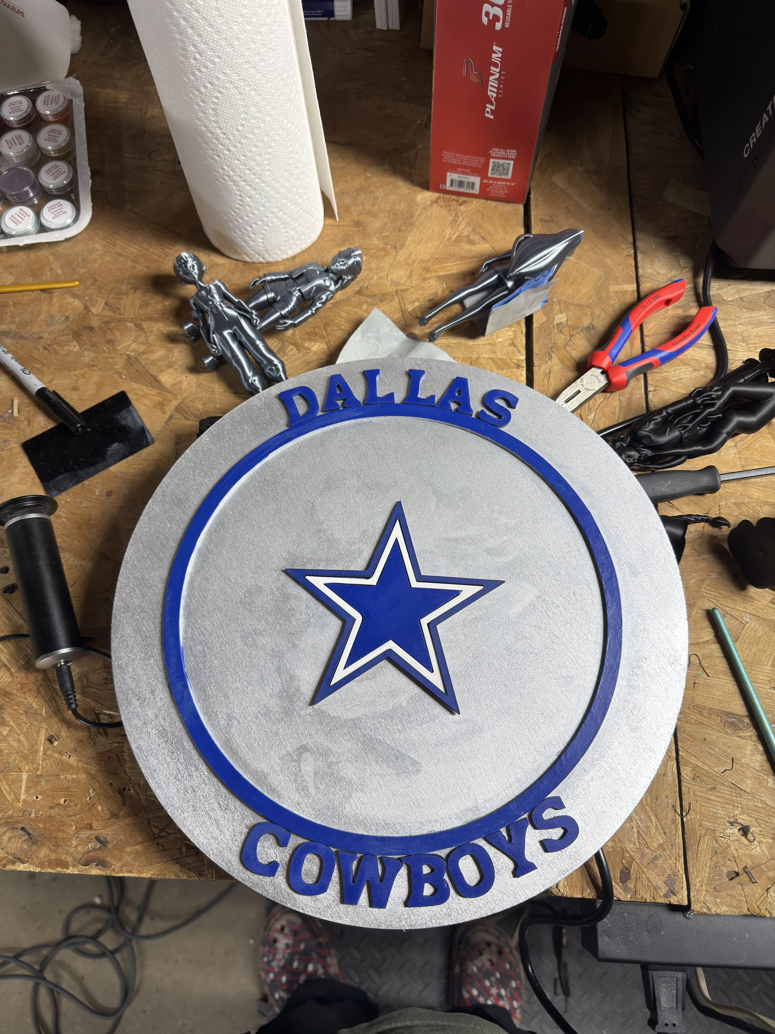 A workbench with a Dallas Cowboys sign, showing a star logo, in the process of being made. Various tools, including pliers, a soldering iron, and figurines, are scattered around.