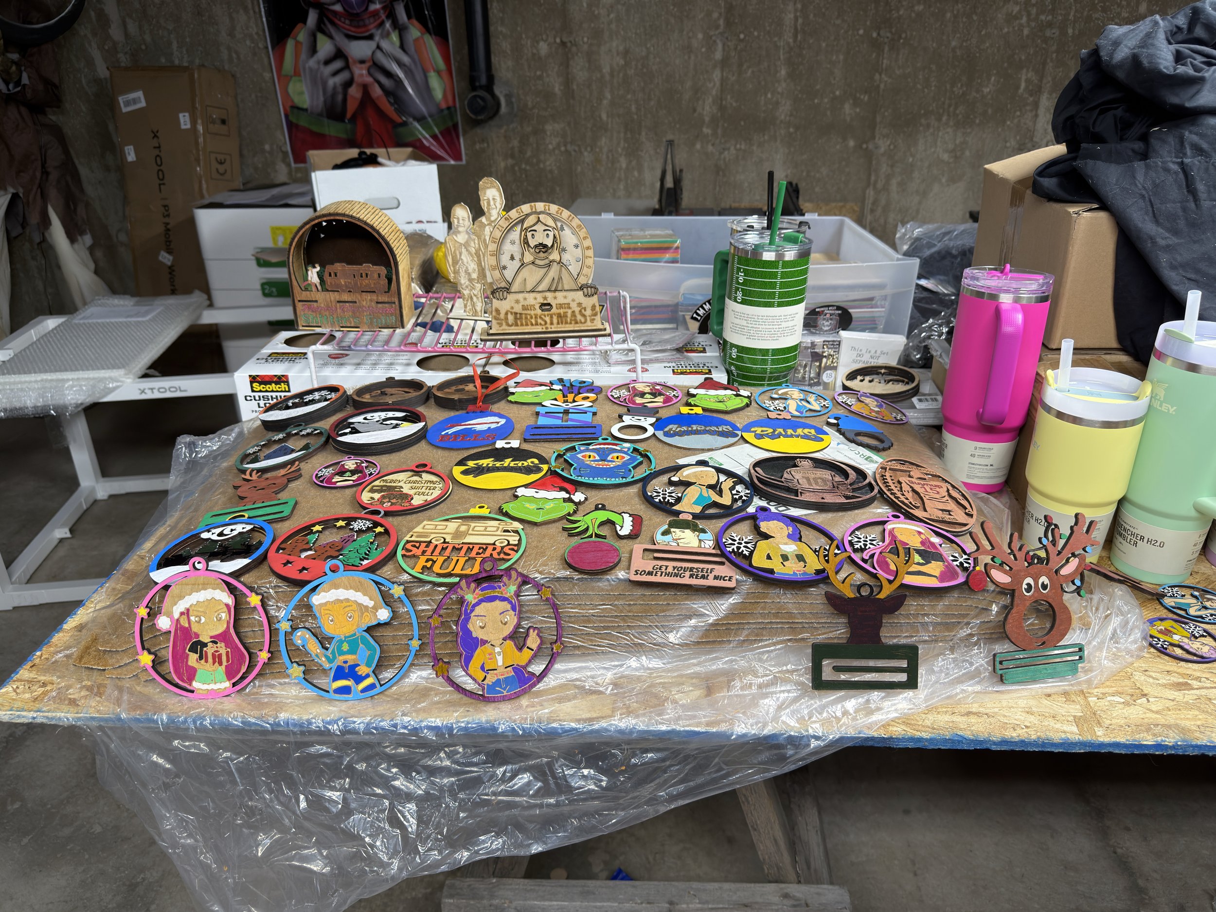 Table displaying various Christmas-themed and decorative patches, keychains, and ornaments with festive designs, alongside reusable cups and bottles, in a workshop or storage area.