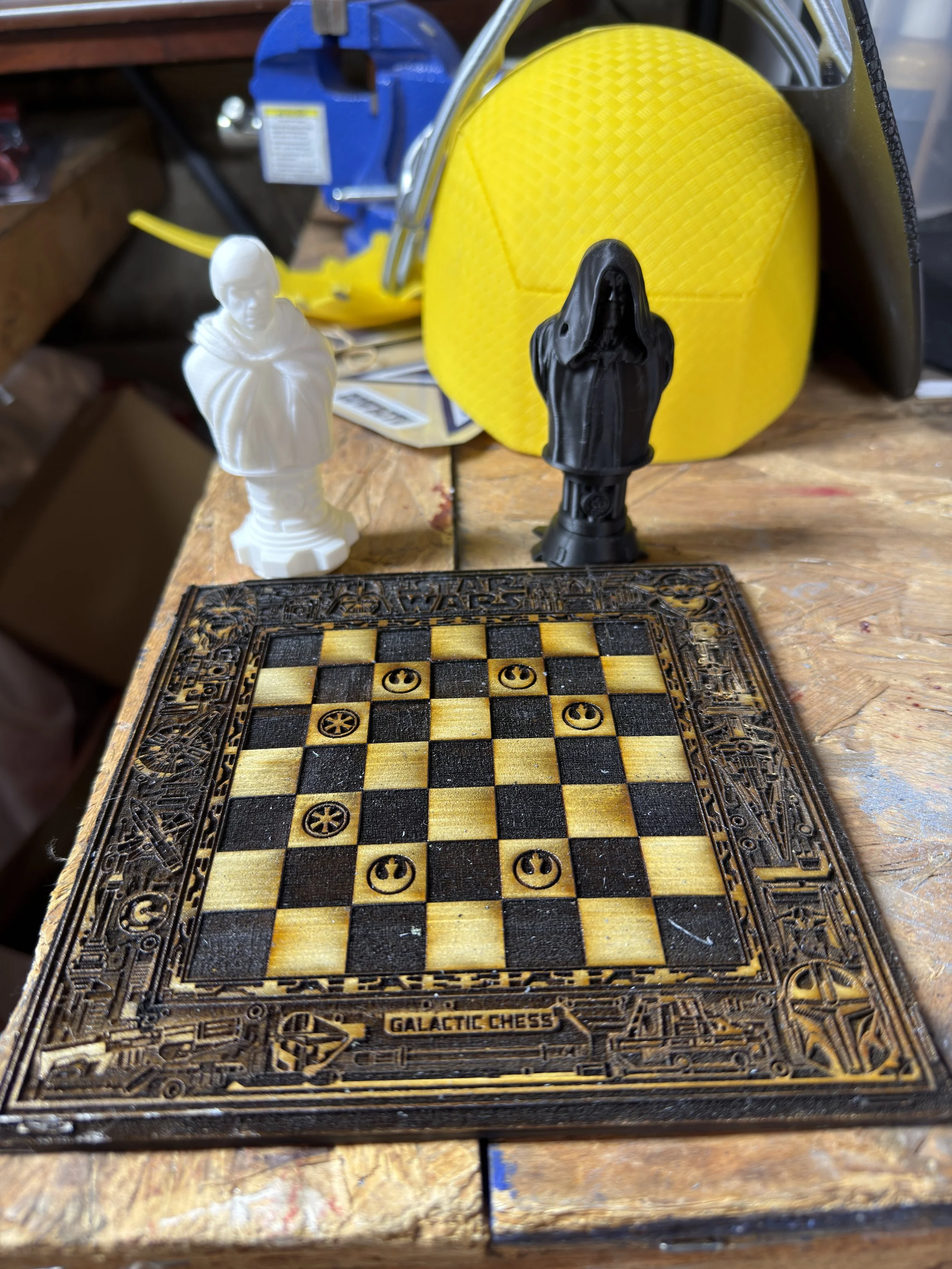 A wooden chessboard with Star Wars-themed chess pieces, including a white Obi-Wan Kenobi and a black Darth Vader, on a workbench surrounded by tools and safety equipment.
