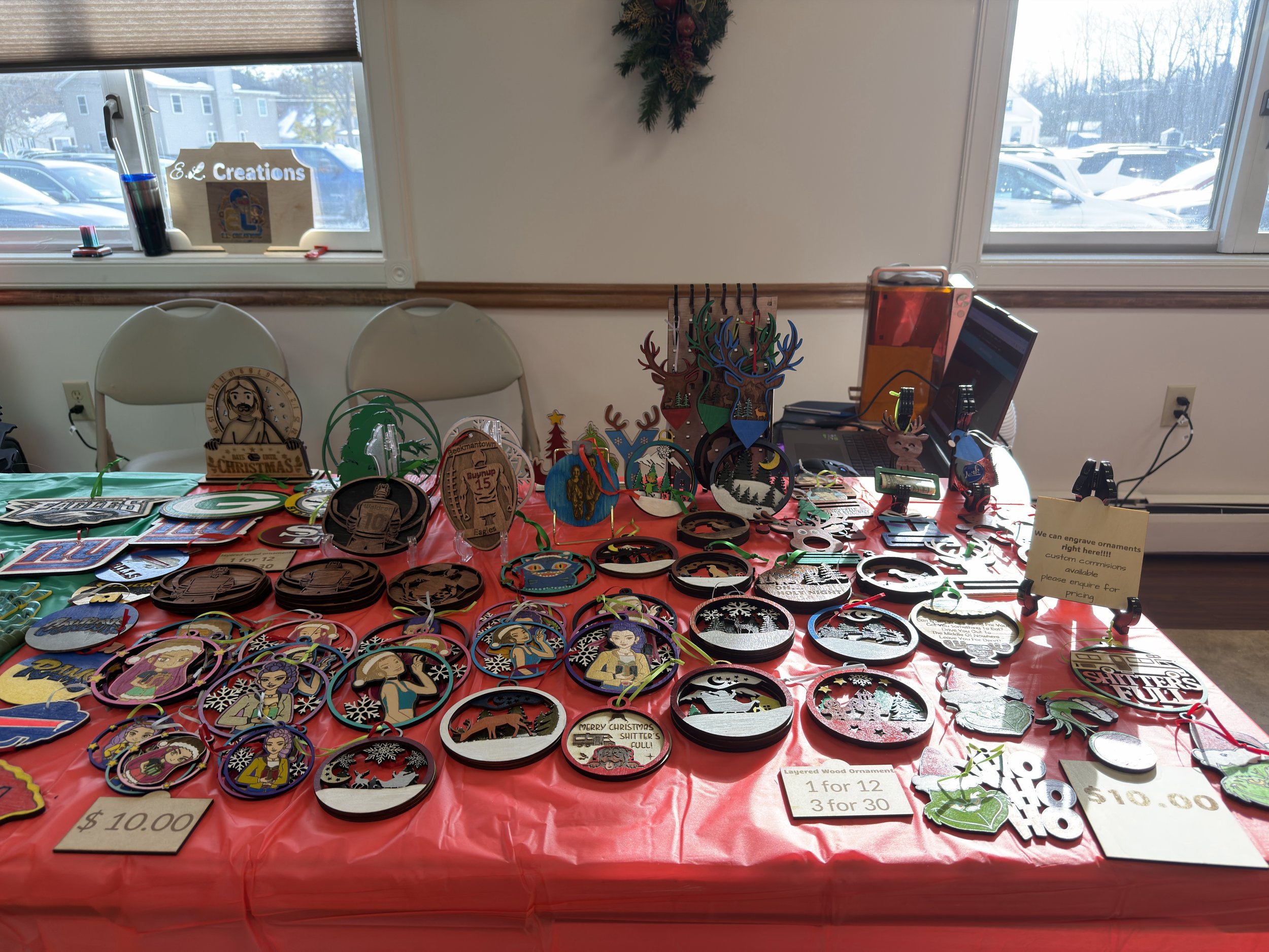 A table displaying various handmade Christmas ornaments for sale, with some priced at $10 each, 3 for $30, and layered wood ornaments at $10.00. The ornaments include reindeer, Santa, winter scenes, and cartoon characters. Behind the table are two chairs, a window, and a computer on the right side.