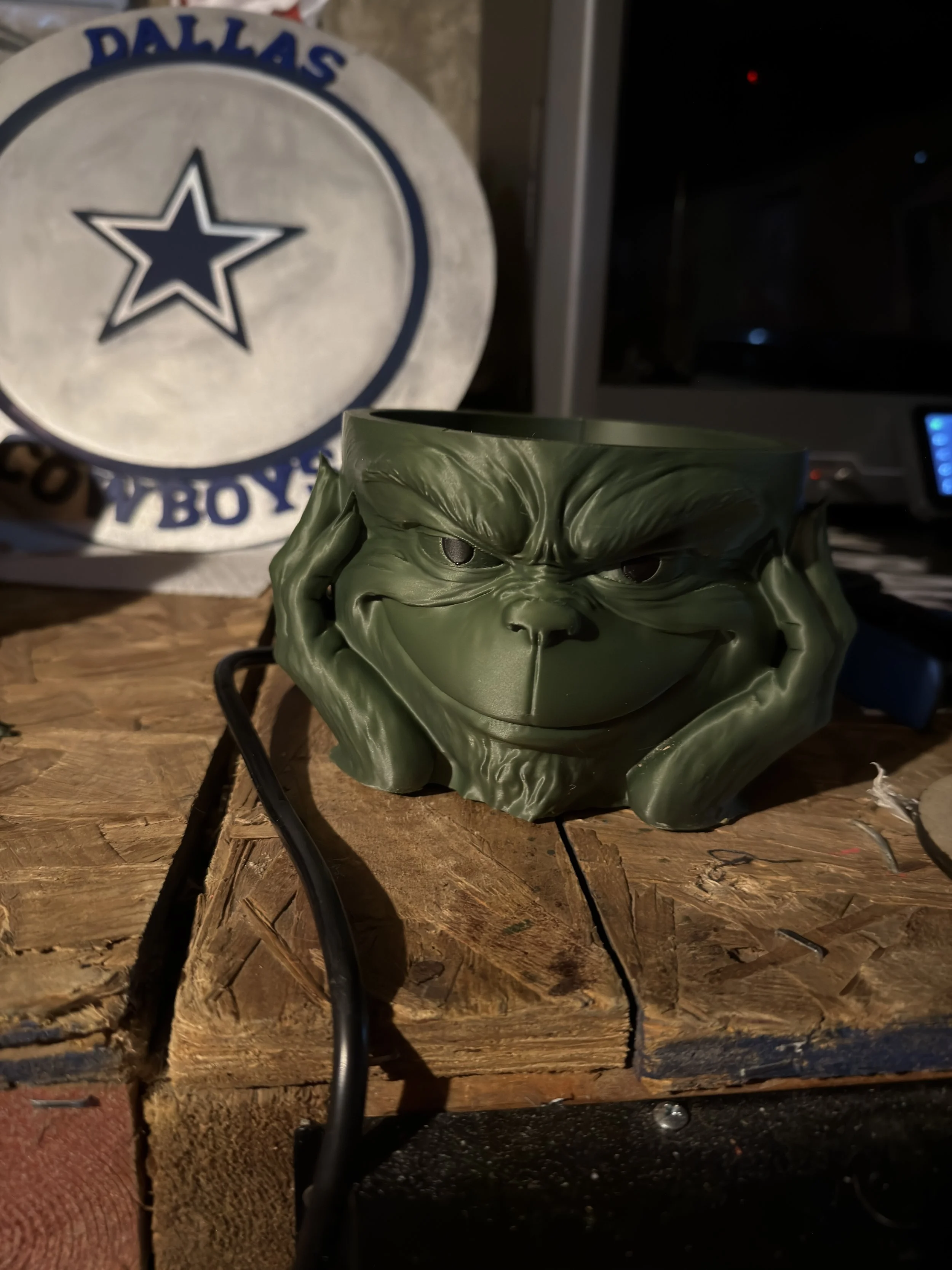 A humorous green mug with a sculpted face of the Grinch smiling, placed on a wooden surface with a Dallas Cowboys decoration in the background.