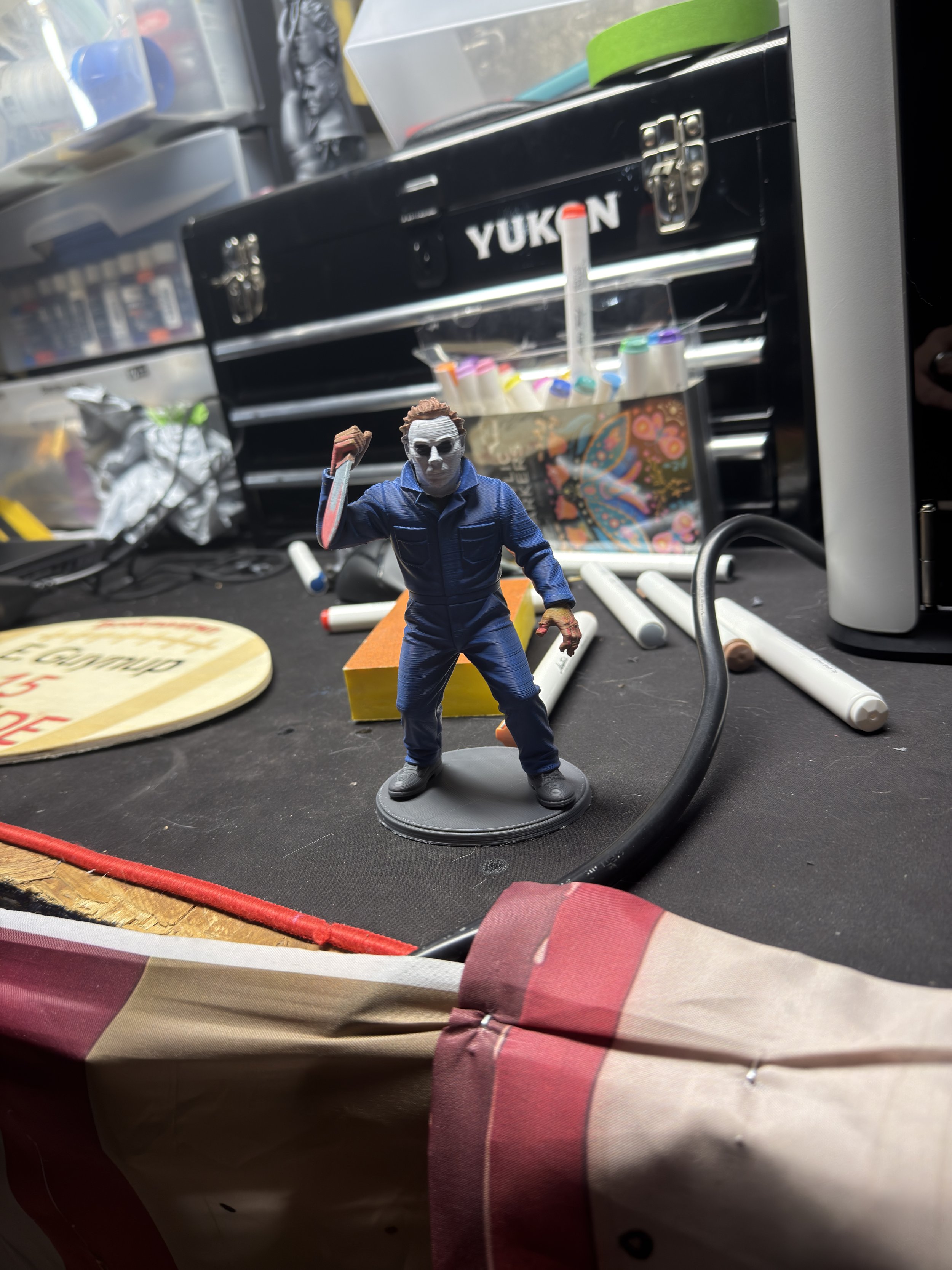 A miniature figurine of Michael Myers from the Halloween movie series is placed on a cluttered workspace with tools and supplies, including markers and a toolbox in the background.