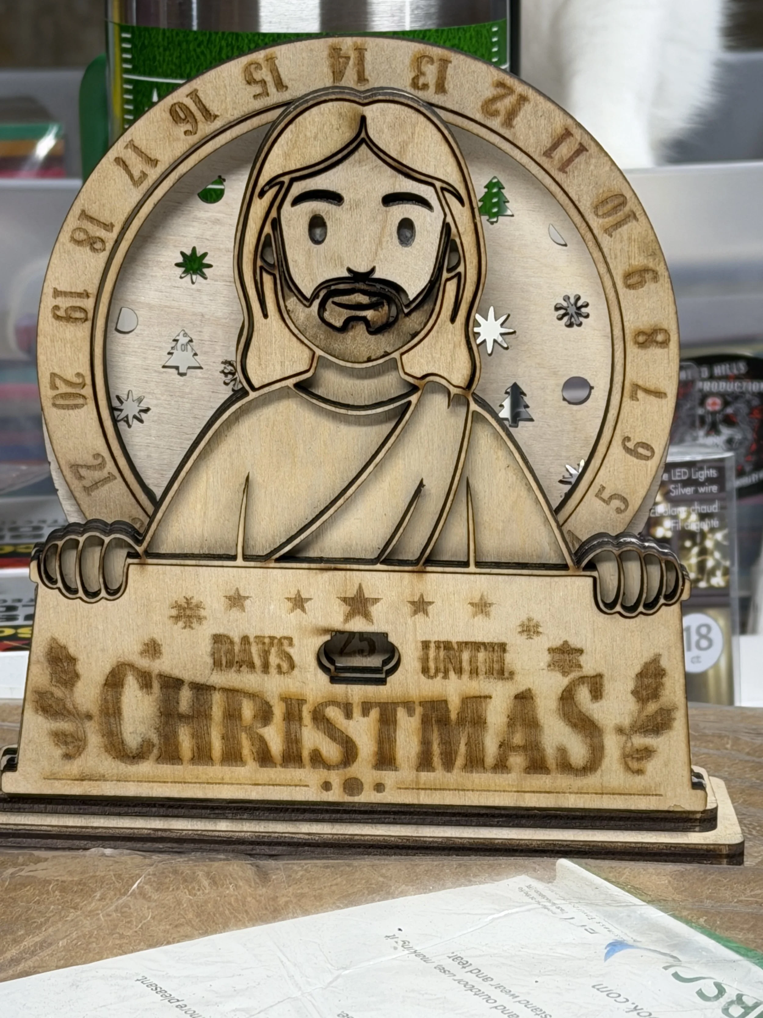 Wooden Christmas countdown clock featuring a Jesus figure holding a sign that reads 'DAYS UNTIL CHRISTMAS,' with a circular calendar and decorative holiday symbols in the background.