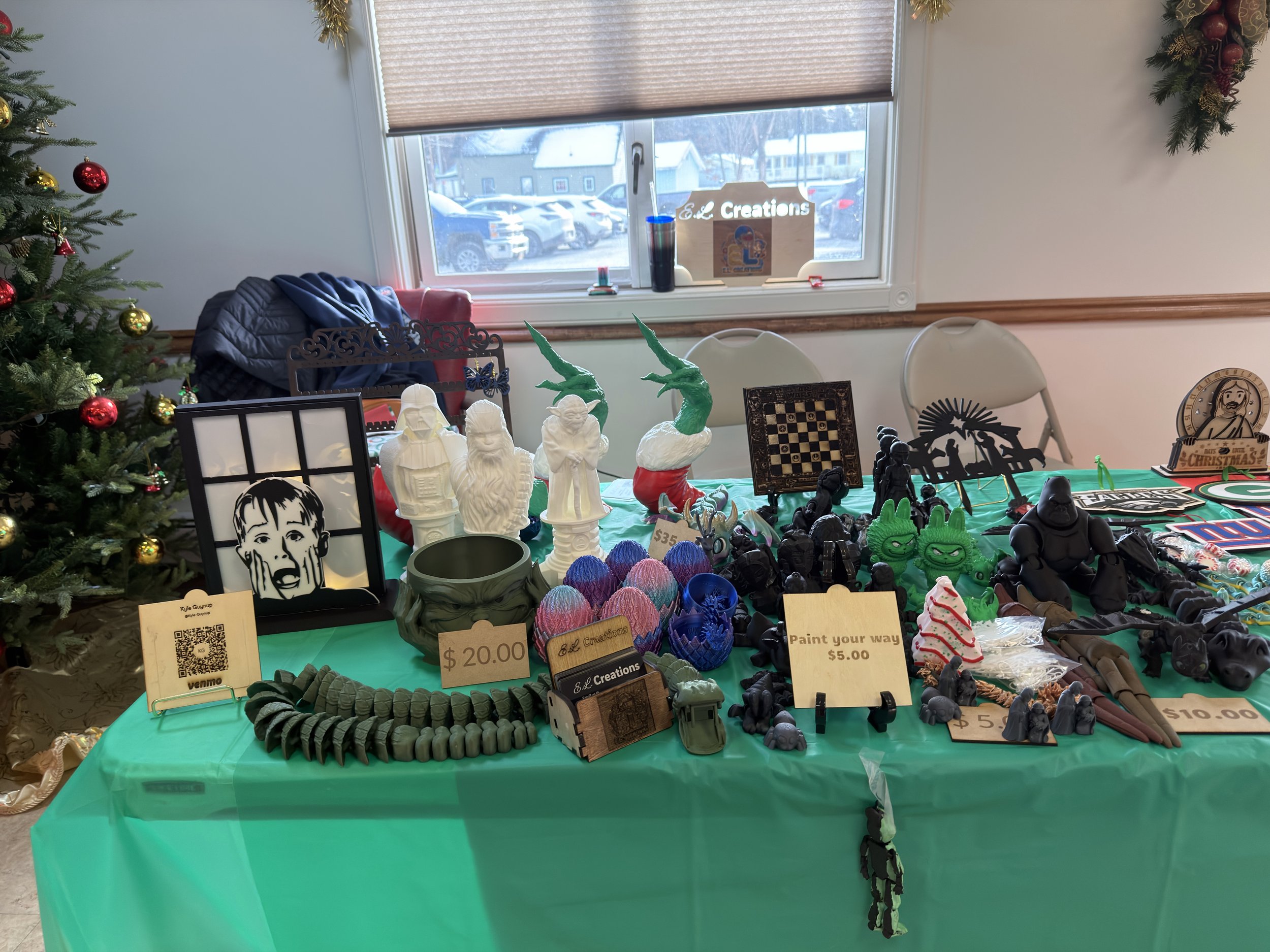 Holiday craft fair display table with various art pieces, figurines, and decorations, including a Christmas tree with ornaments, a window with a sign, and a green tablecloth.