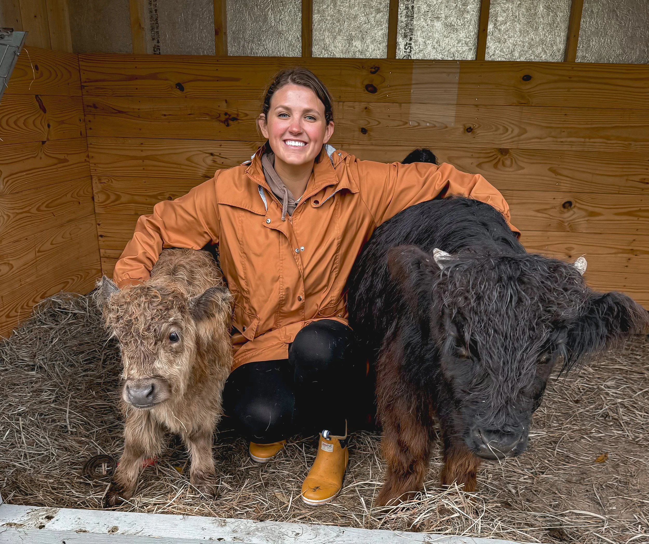 A woman kneeling inside a barn with a small calf and a large black dog. The woman is wearing an orange rain jacket and yellow rain boots, smiling at the camera. The small calf is sitting on the straw-covered ground, and the black dog is standing beside the woman.