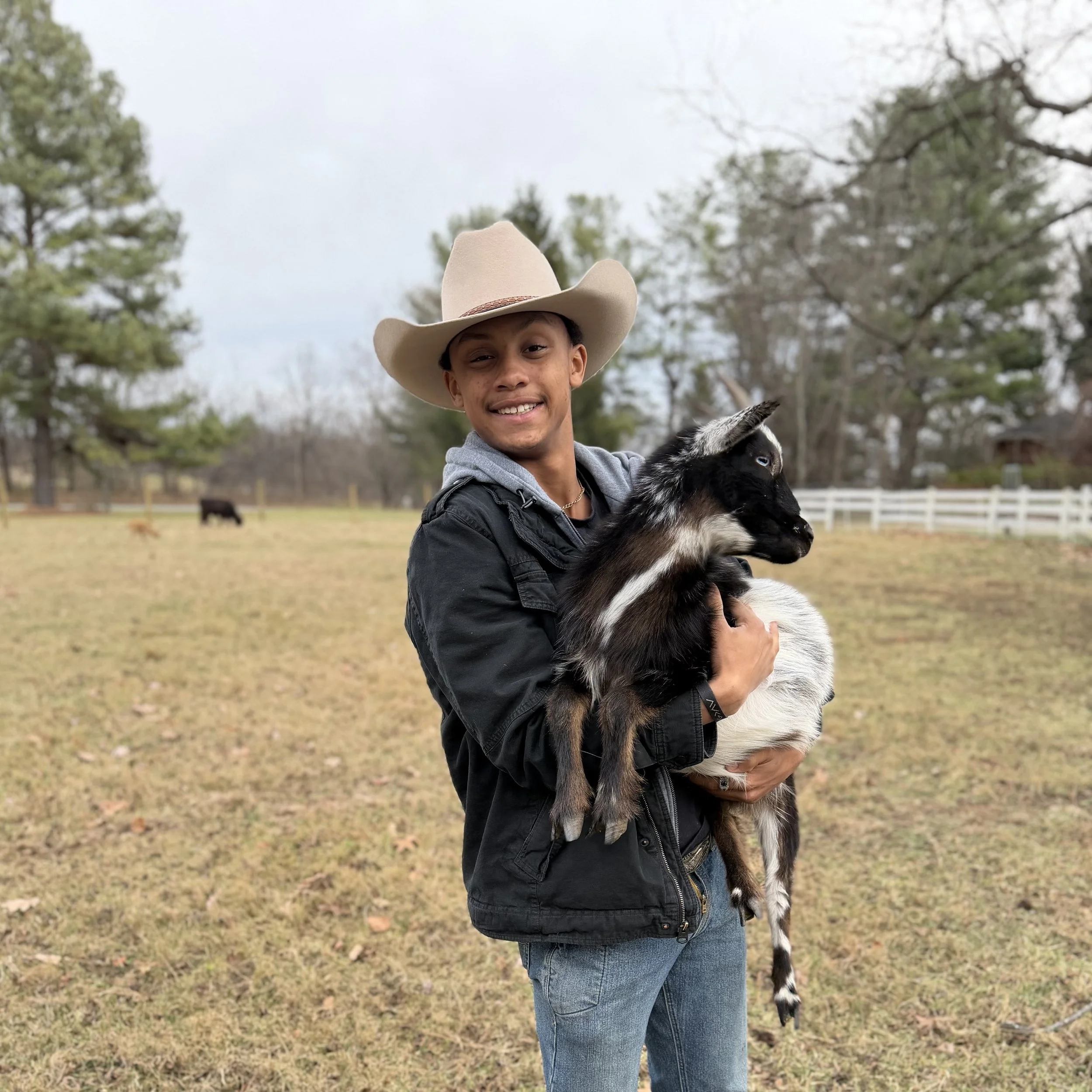 A young man wearing a cowboy hat and a black jacket holding a baby goat outdoors in a grassy area with trees and a white fence in the background, smiling.