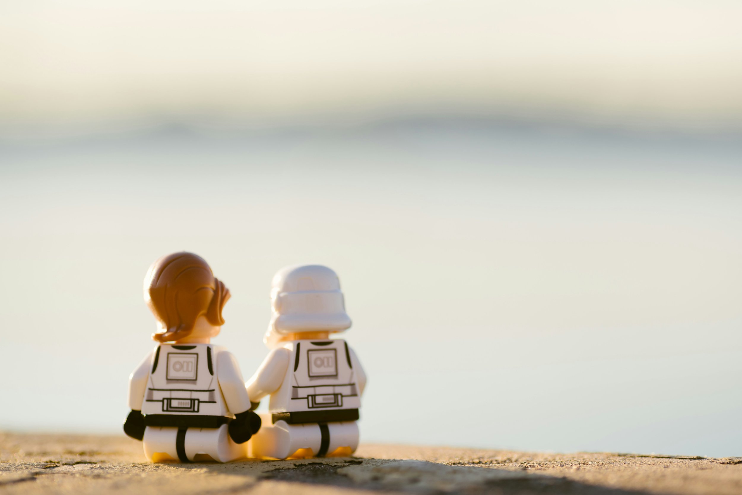 LEGO figures of a woman and a person in a stormtrooper helmet sitting on a surface, with a blurred background of water and sky.