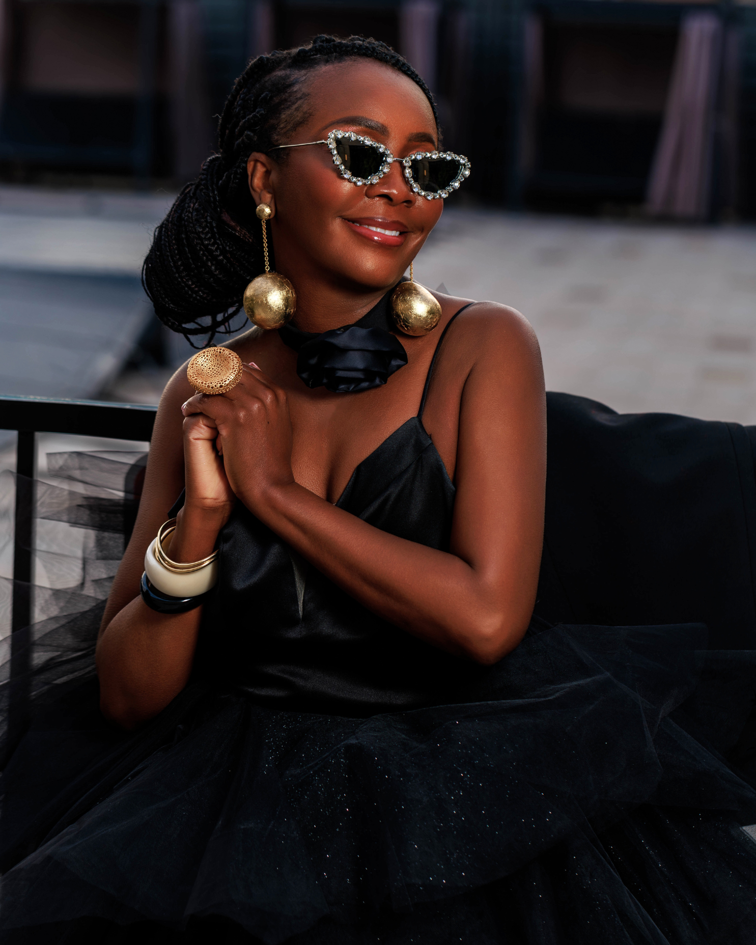 A woman with dark skin wearing stylish sunglasses with rhinestones, large gold earrings, a black satin dress with thin straps, and layered bangles, sitting outdoors on a black chair, smiling with arms crossed.