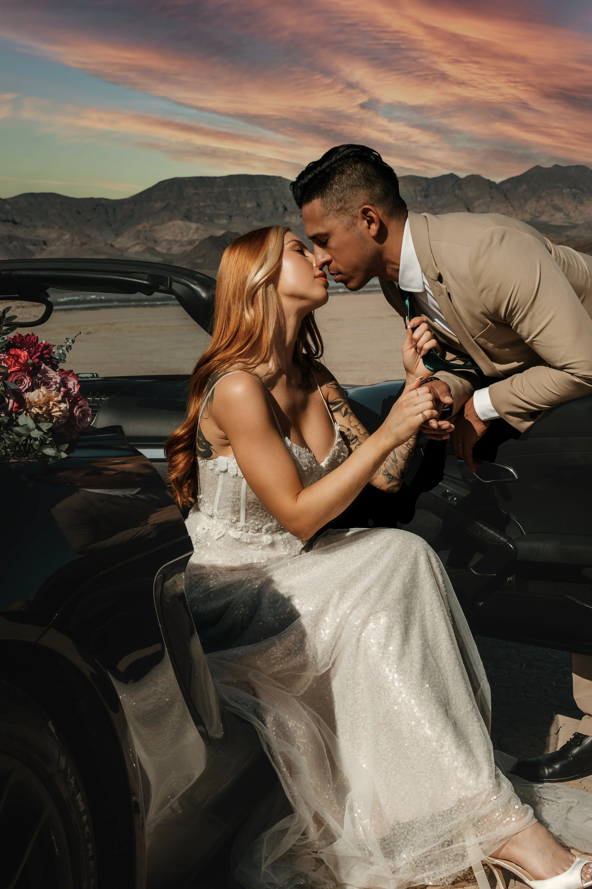 A wedding couple sharing a kiss near a black car decorated with flowers, against a desert landscape at sunset.
