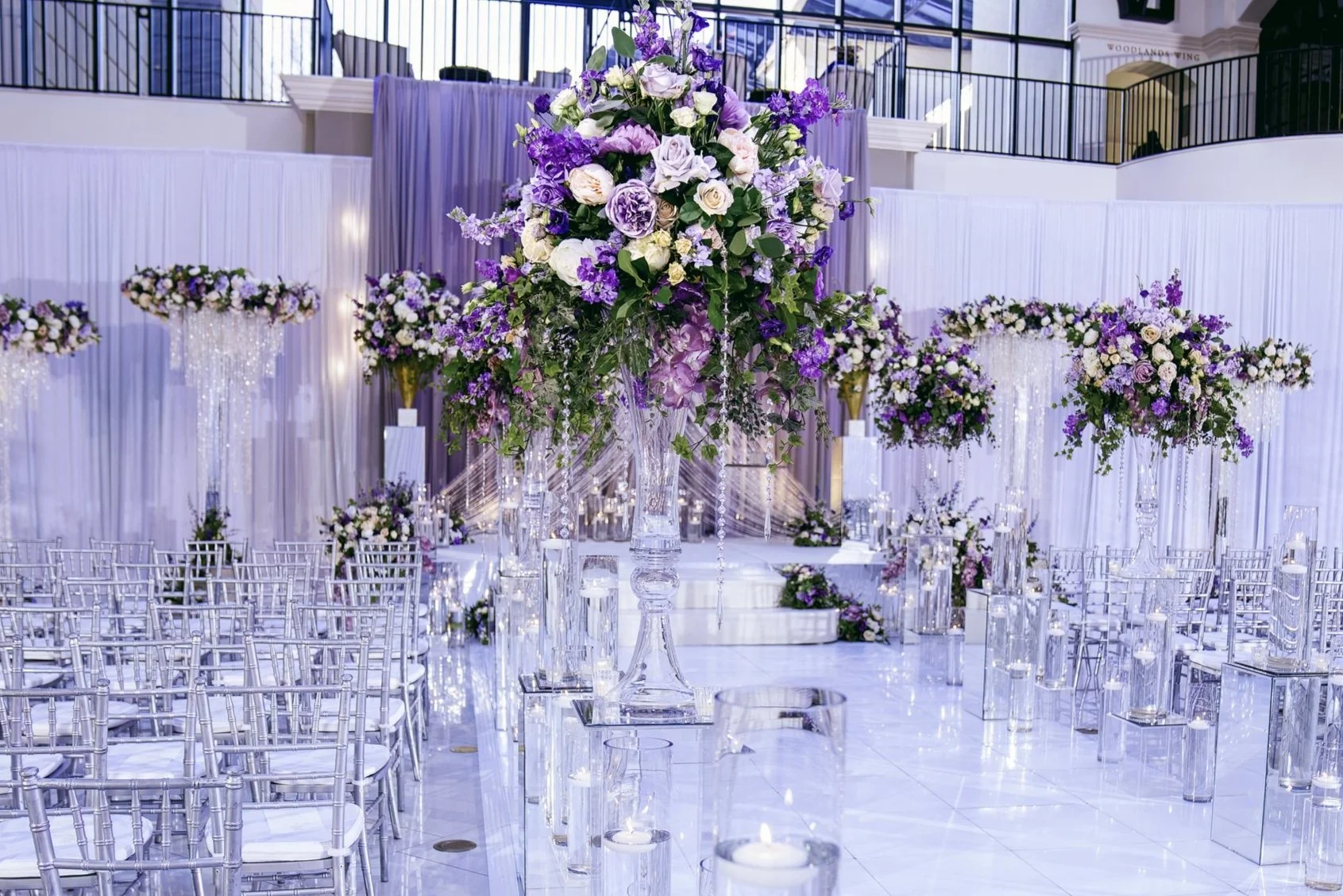 A beautifully decorated indoor wedding venue with a large floral centerpiece and smaller floral arrangements, featuring purple and white flowers, with white chairs arranged for guests and candle displays.