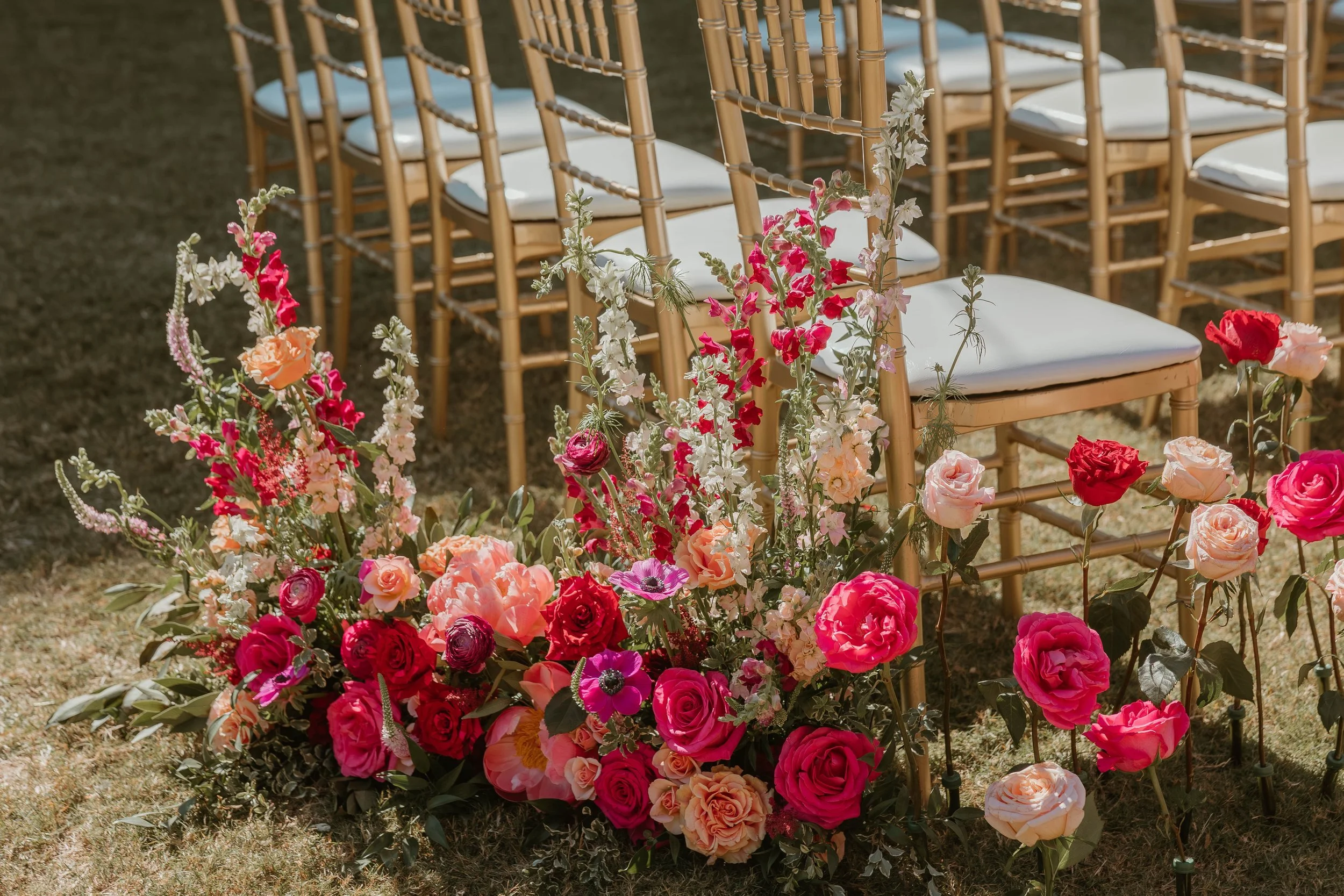 Rows of gold-colored chairs with white cushions arranged outdoors for a wedding, with a large floral arrangement of pink, red, white, and peach flowers on the grass in front of the chairs. At Chateau Elan